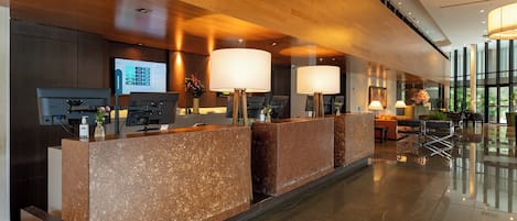 A hotel reception area with a modern design, featuring a reception desk, a digital screen, and a seating area with a view of the outdoors.