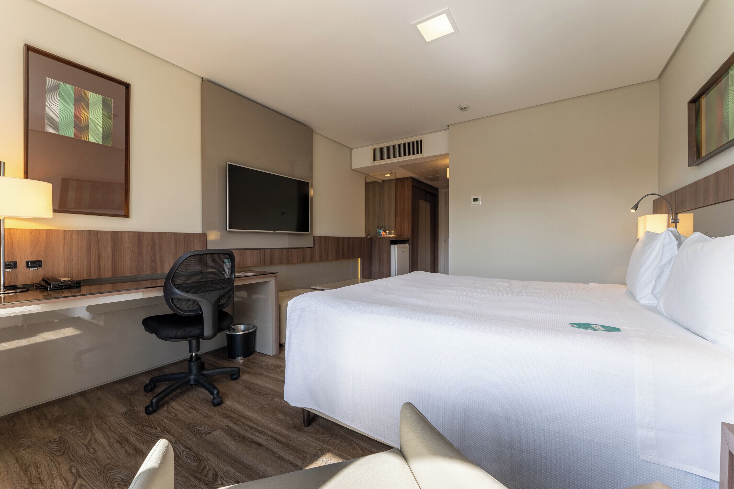 A hotel room with a bed, desk, chair, TV, and a small table with a lamp.