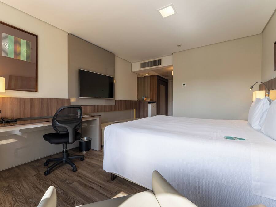 A hotel room with a bed, desk, chair, TV, and a small table with a lamp.