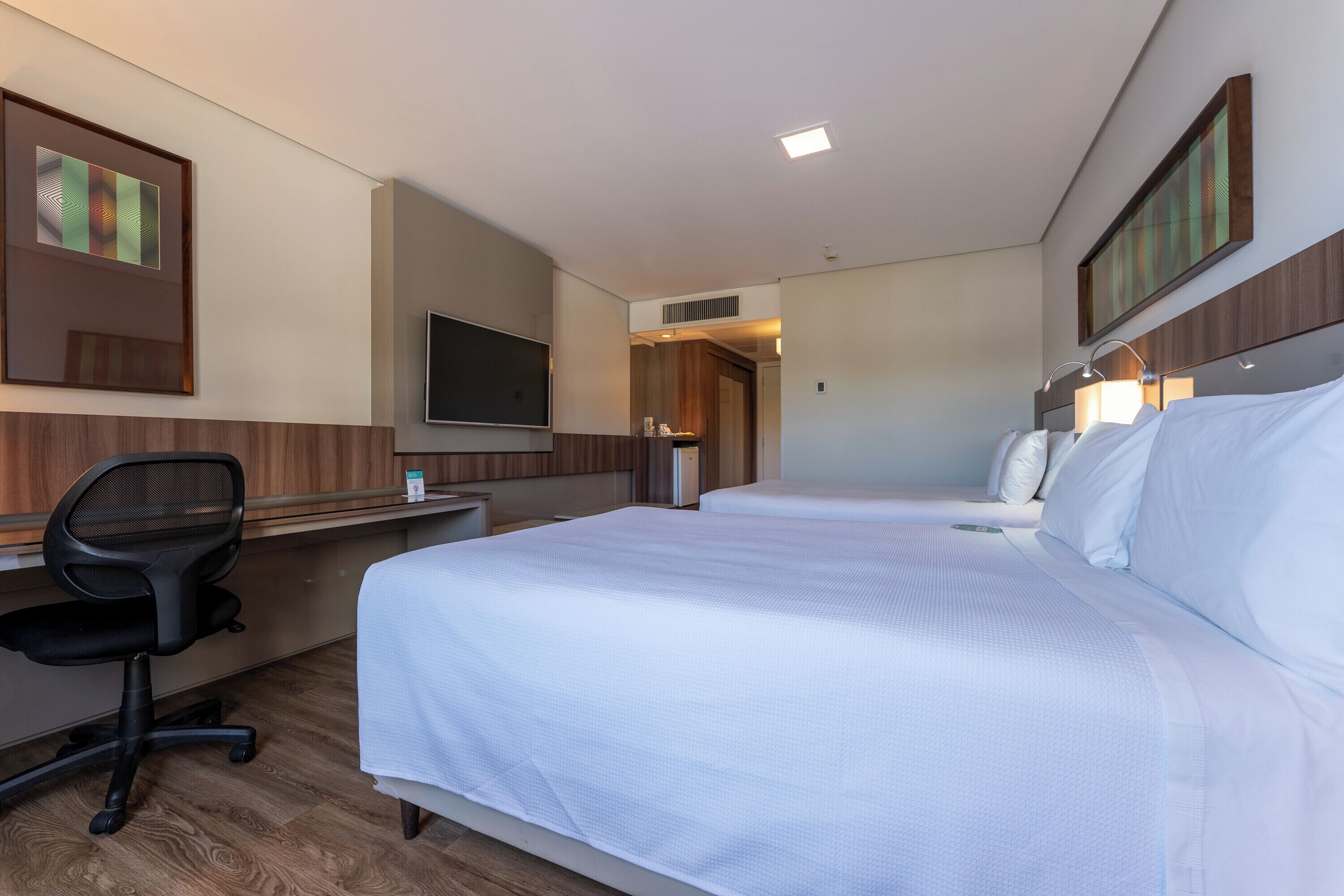 Habitación de hotel con dos camas, un escritorio con silla, televisión y obras de arte en las paredes.