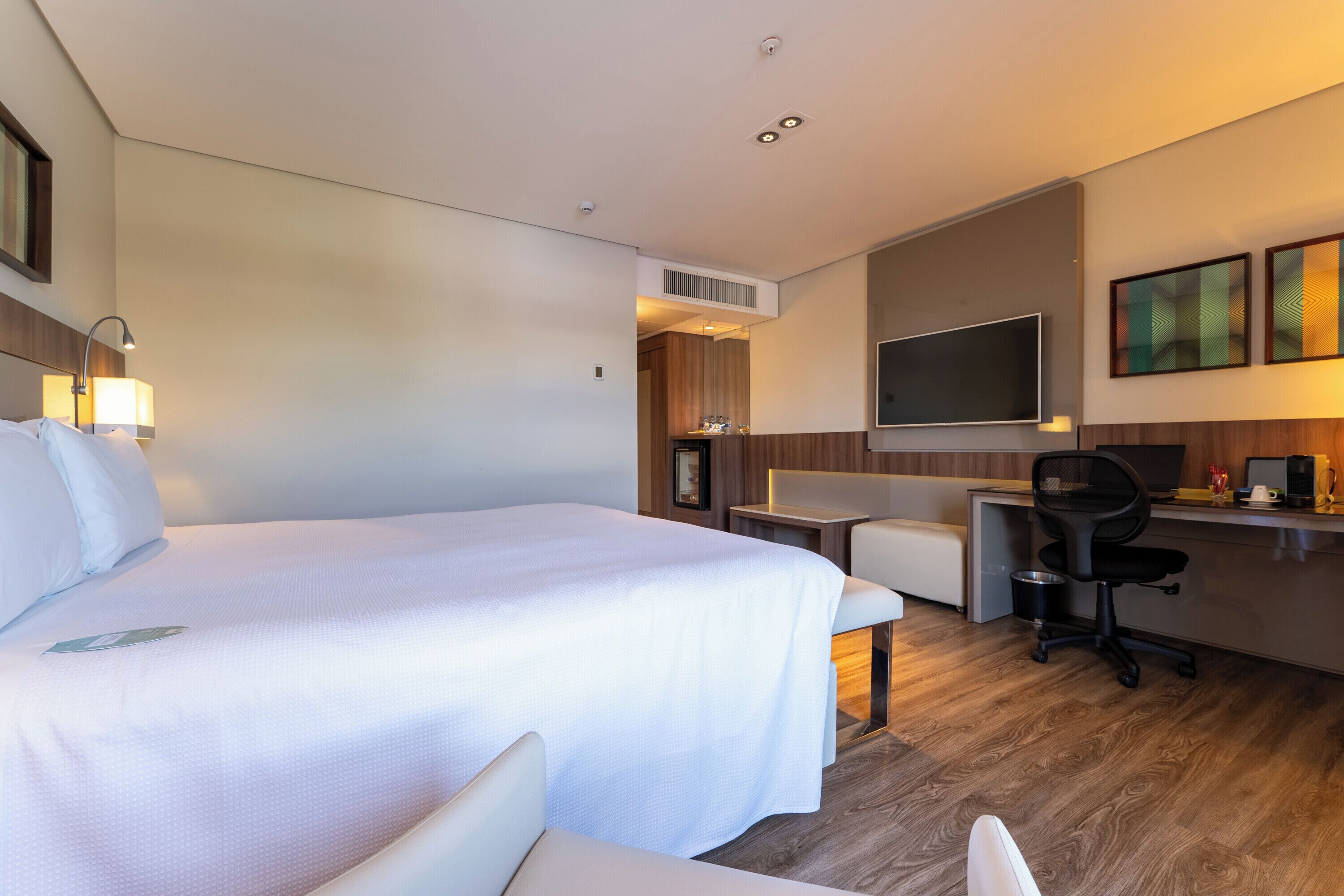 A hotel room with a large bed, a desk, a chair, a TV, and a small kitchenette.