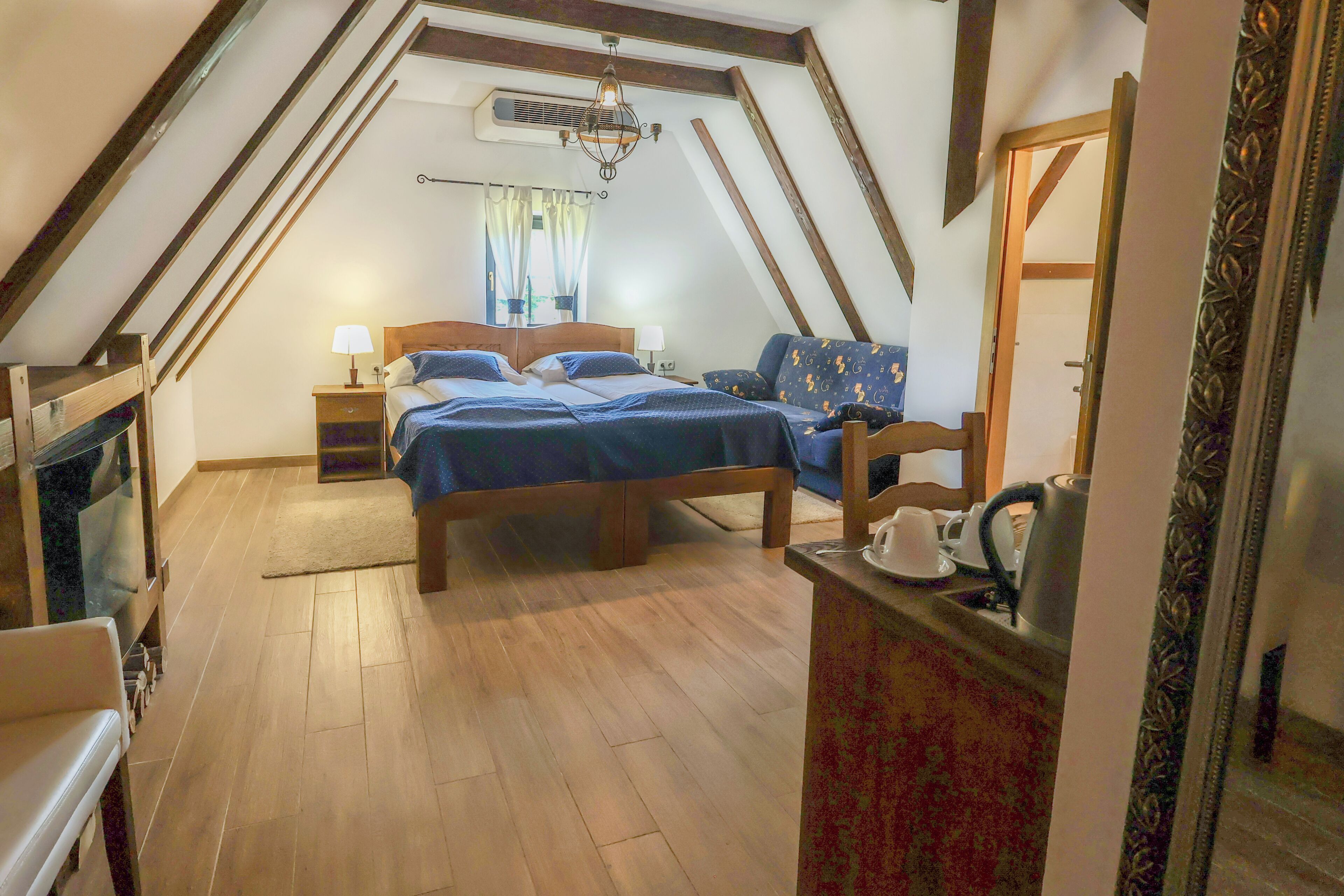 A bedroom with a lofted ceiling, wooden flooring, a bed with blue bedding, a bedside table with a lamp, a dresser, and a mirror.