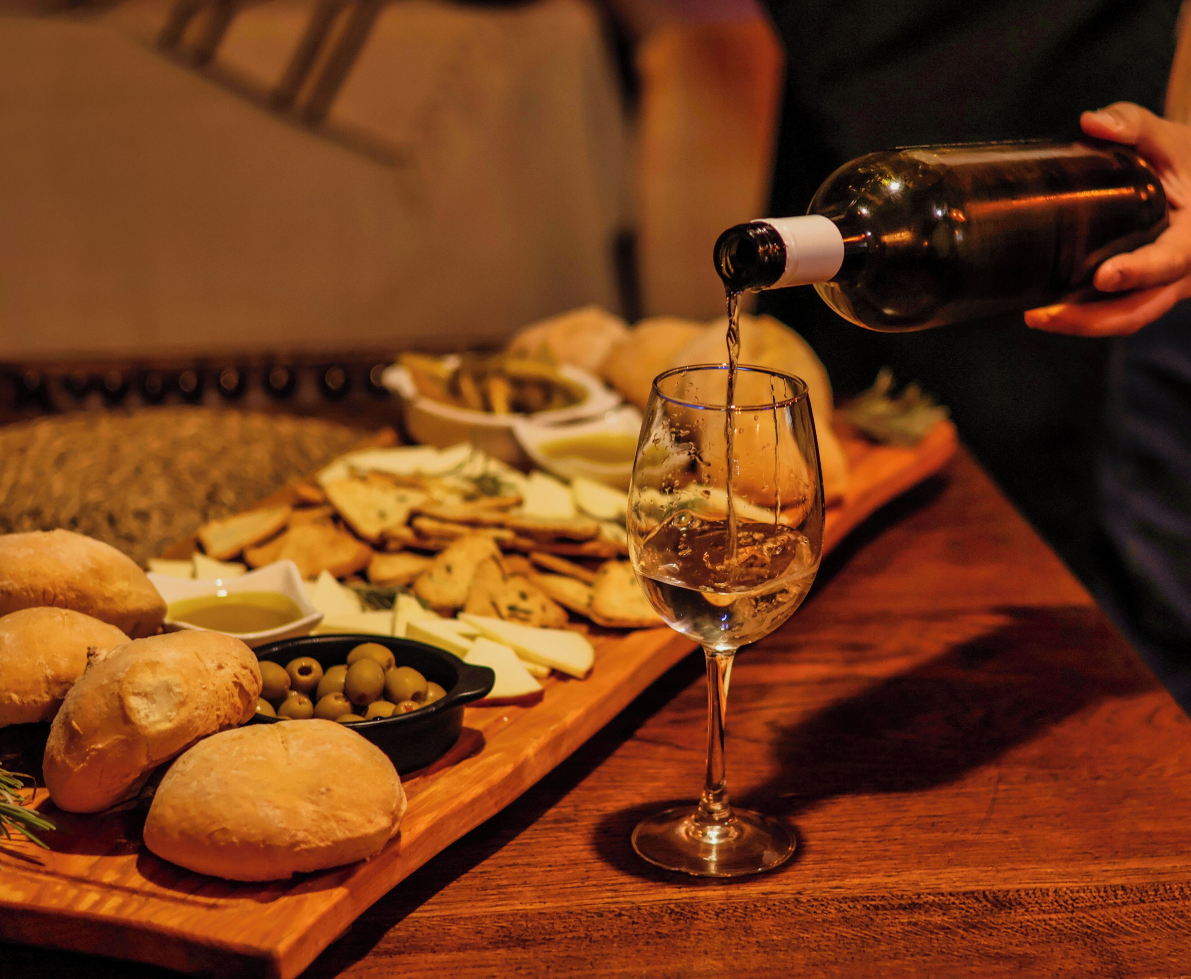 A wooden table with a glass of white wine, a bottle being poured, and a variety of snacks.