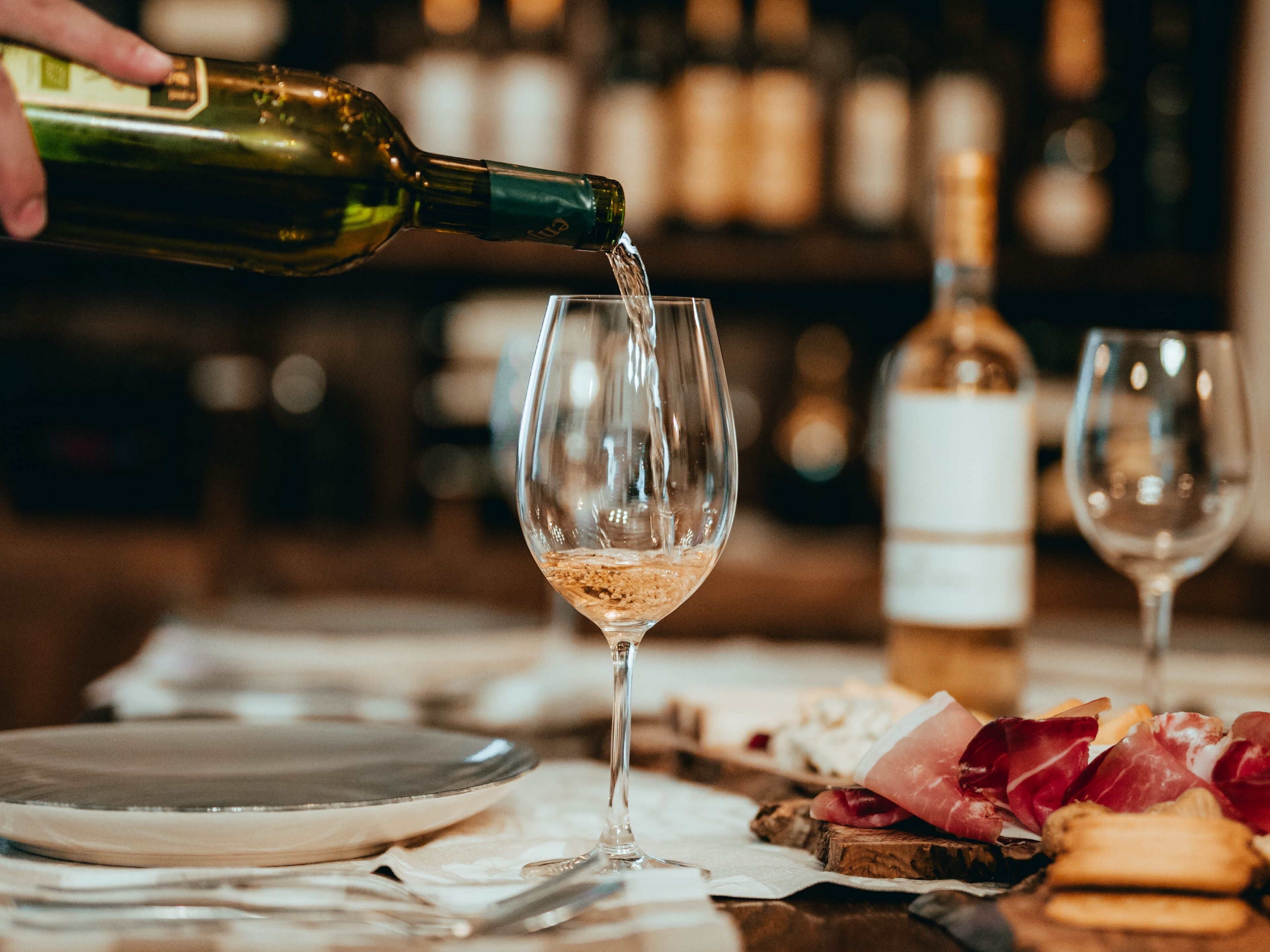 A wine bottle being poured into a glass, with a selection of cheeses and cured meats on the table.