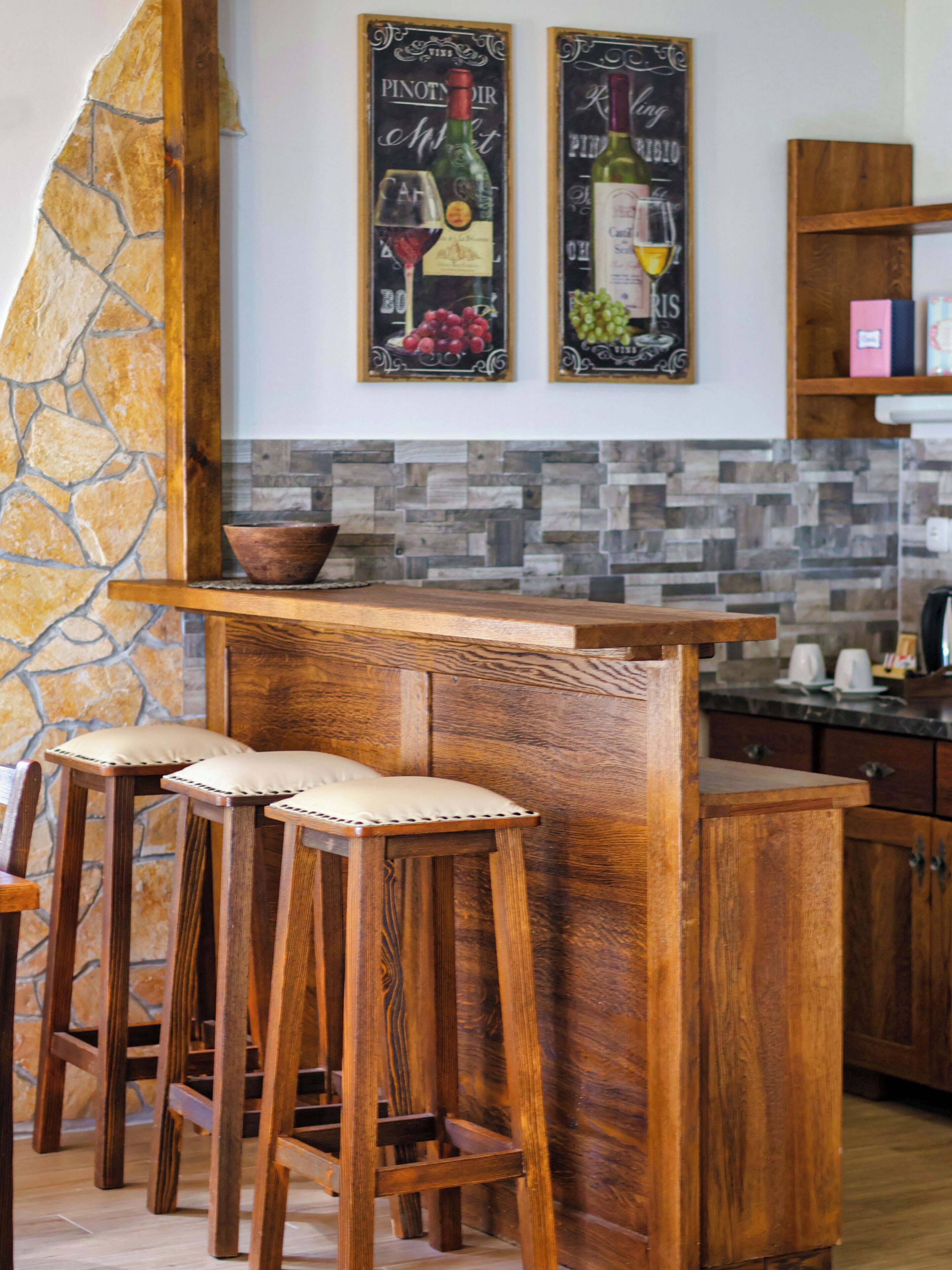 A kitchen with a stone accent wall, wooden bar stools, and two framed wine posters.