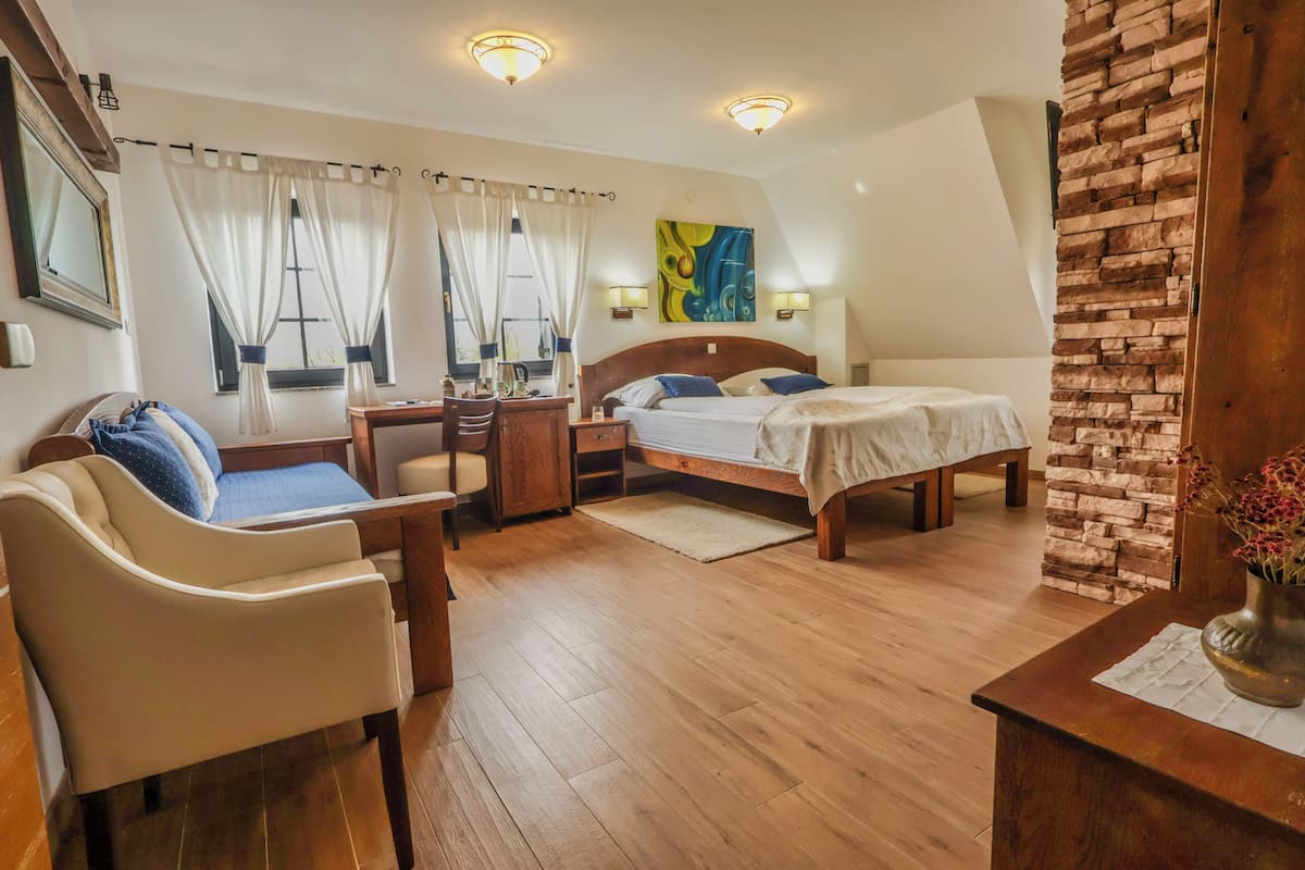 A hotel room with a bed, a desk, a chair, a small table, curtains, a painting, and a brick wall.