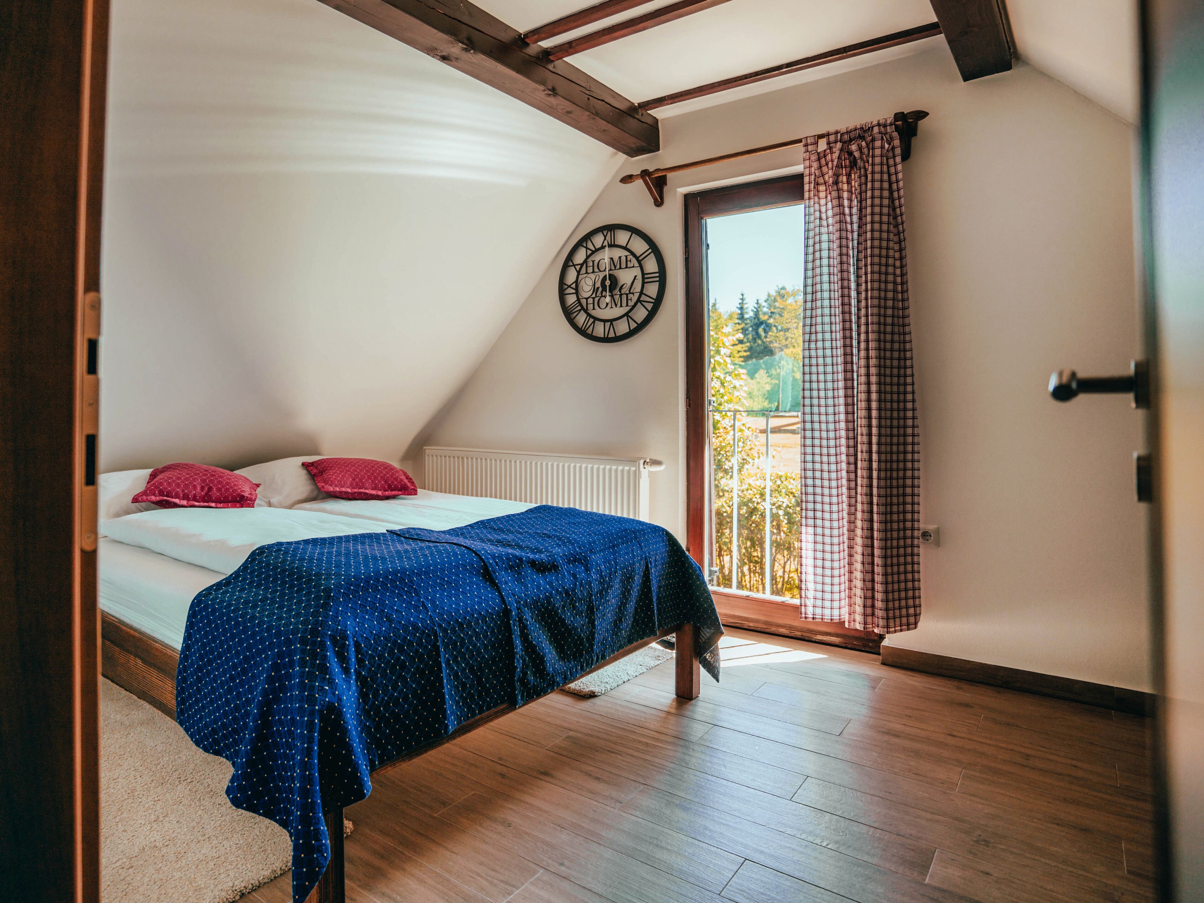 A bedroom with a sloped ceiling, a bed with a blue bedspread, two pillows, a wall clock, and a window with curtains.