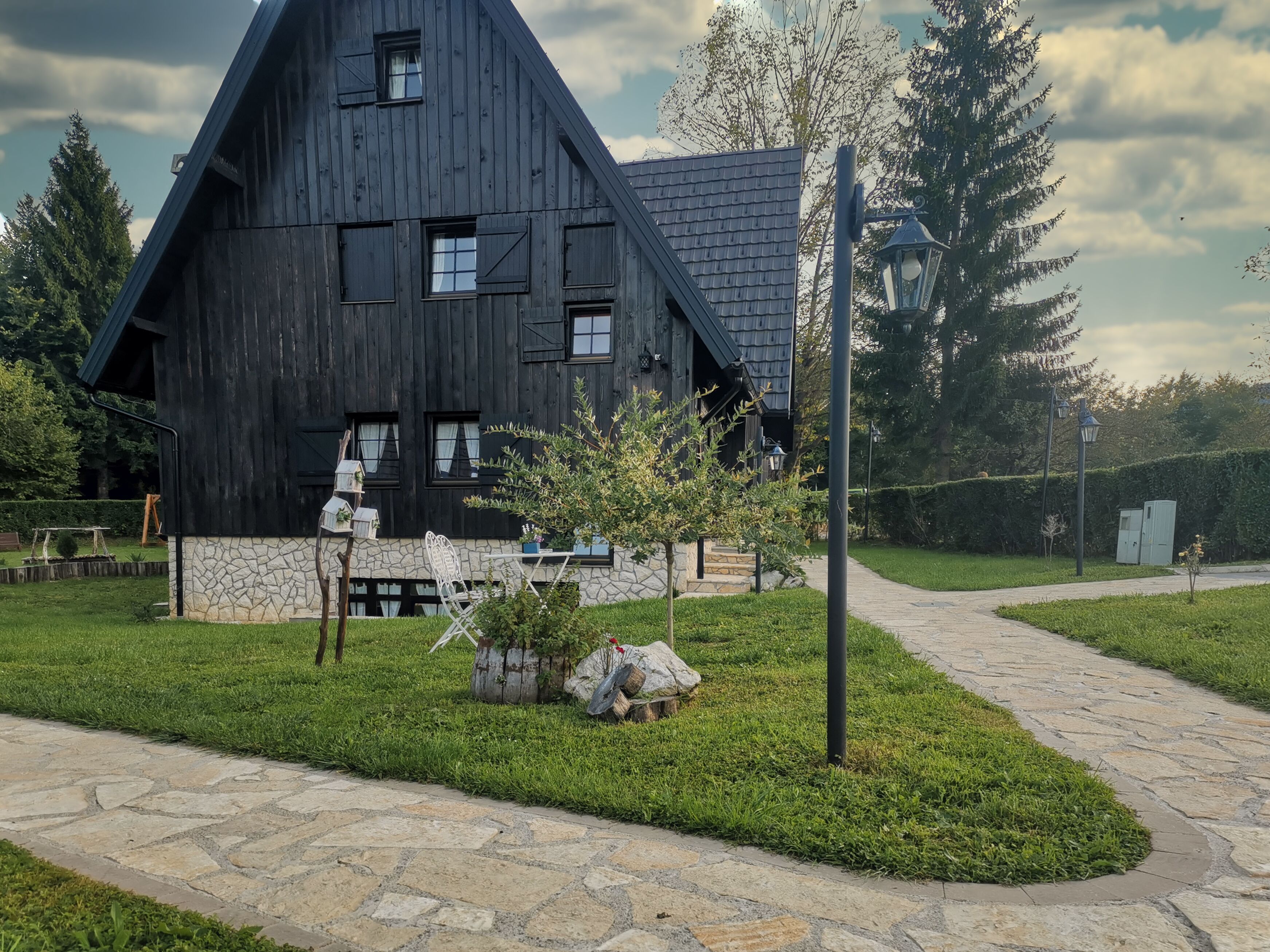 Een rustiek houten gebouw met een stenen fundering, een verhard pad en een kleine tuin met een bankje en plantenpotten.