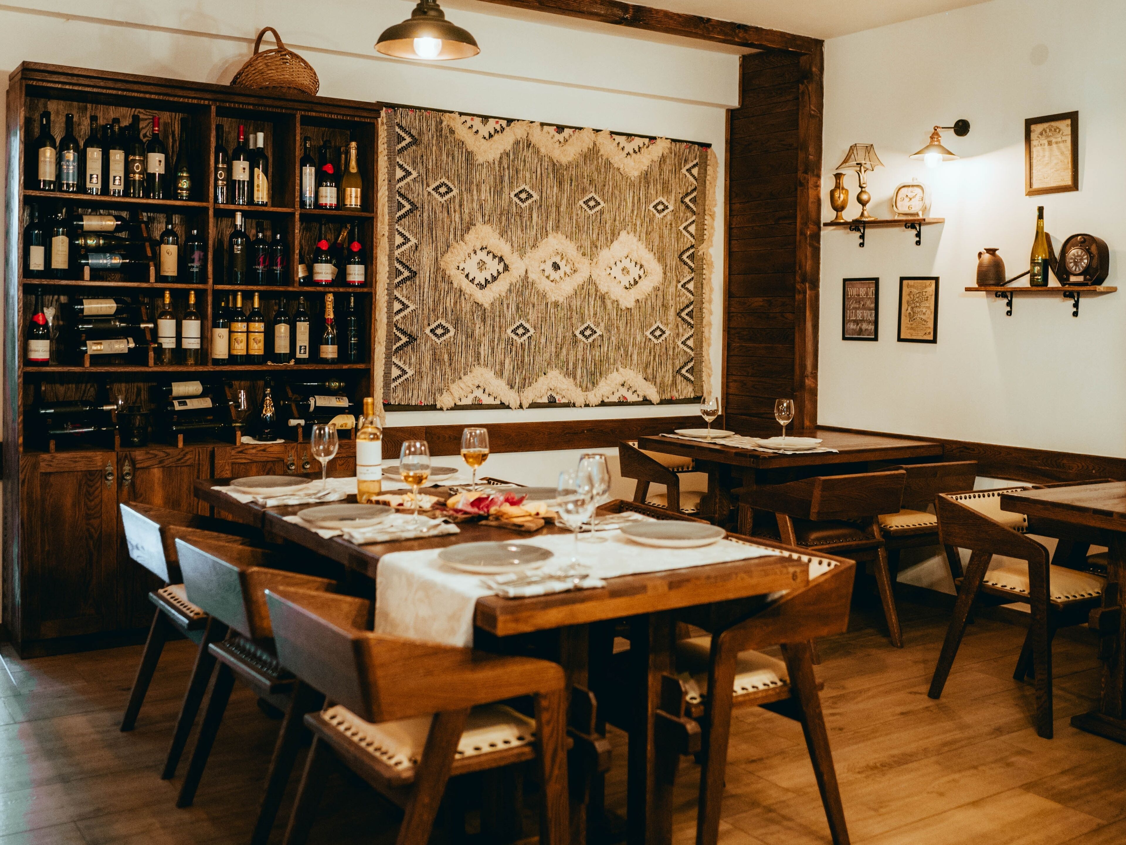 A restaurant interior with wooden furniture, a wine rack, and a patterned wall hanging.