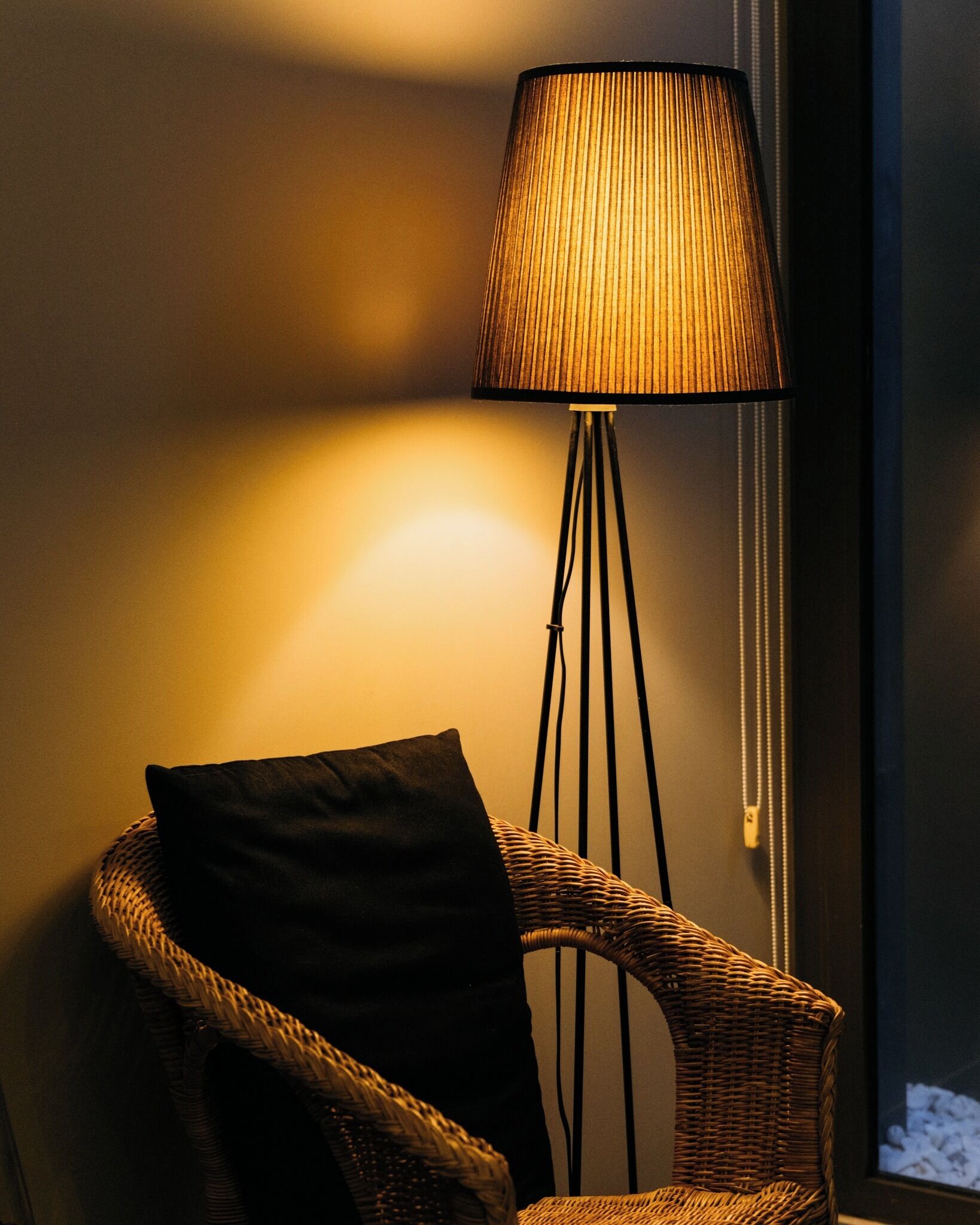 Ein Korbstuhl mit schwarzem Sitzkissen, eine Stehlampe mit geripptem Lampenschirm und eine Wandleuchte.