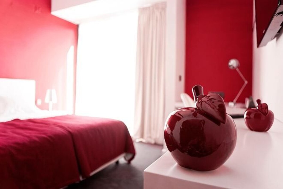A hotel room with a bed, a white nightstand, and a red wall.