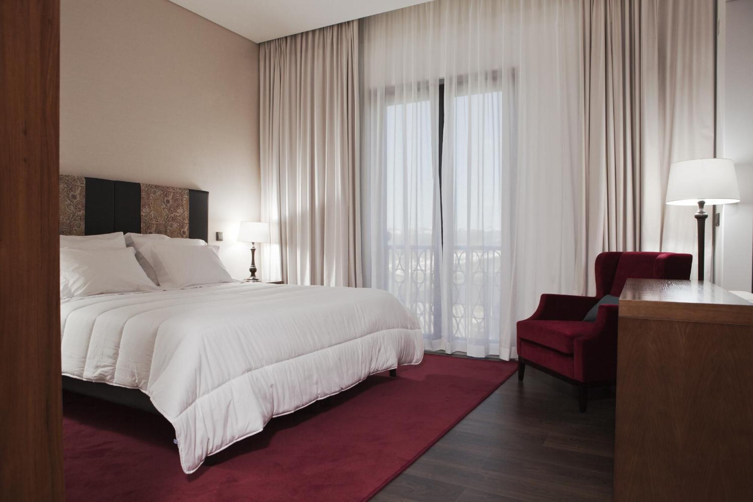 A hotel room with a large bed, a red chair, a desk with a lamp, and sheer curtains.