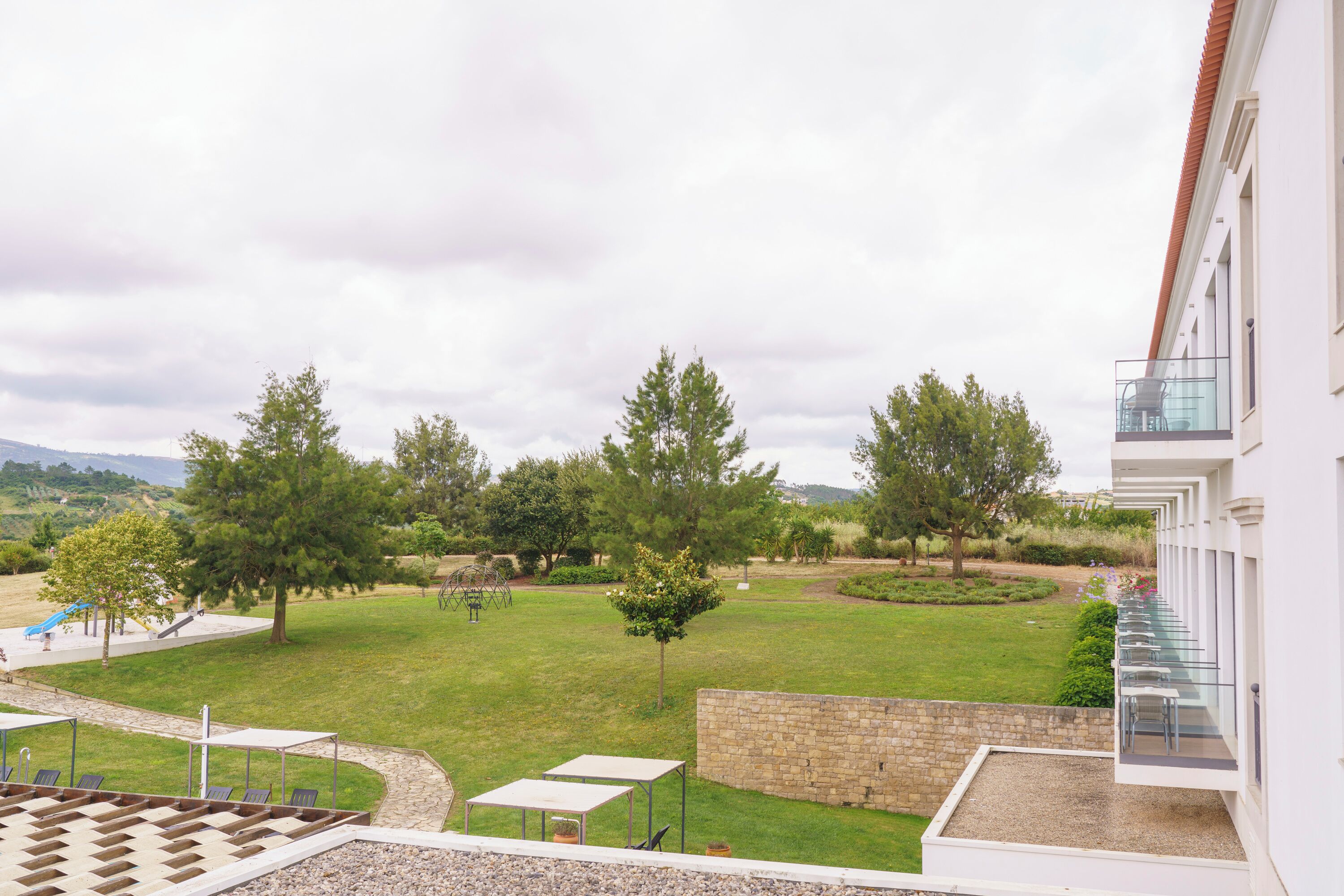 A view of a courtyard with a grassy lawn, trees, and a playground. There are outdoor seating areas and a stone wall.