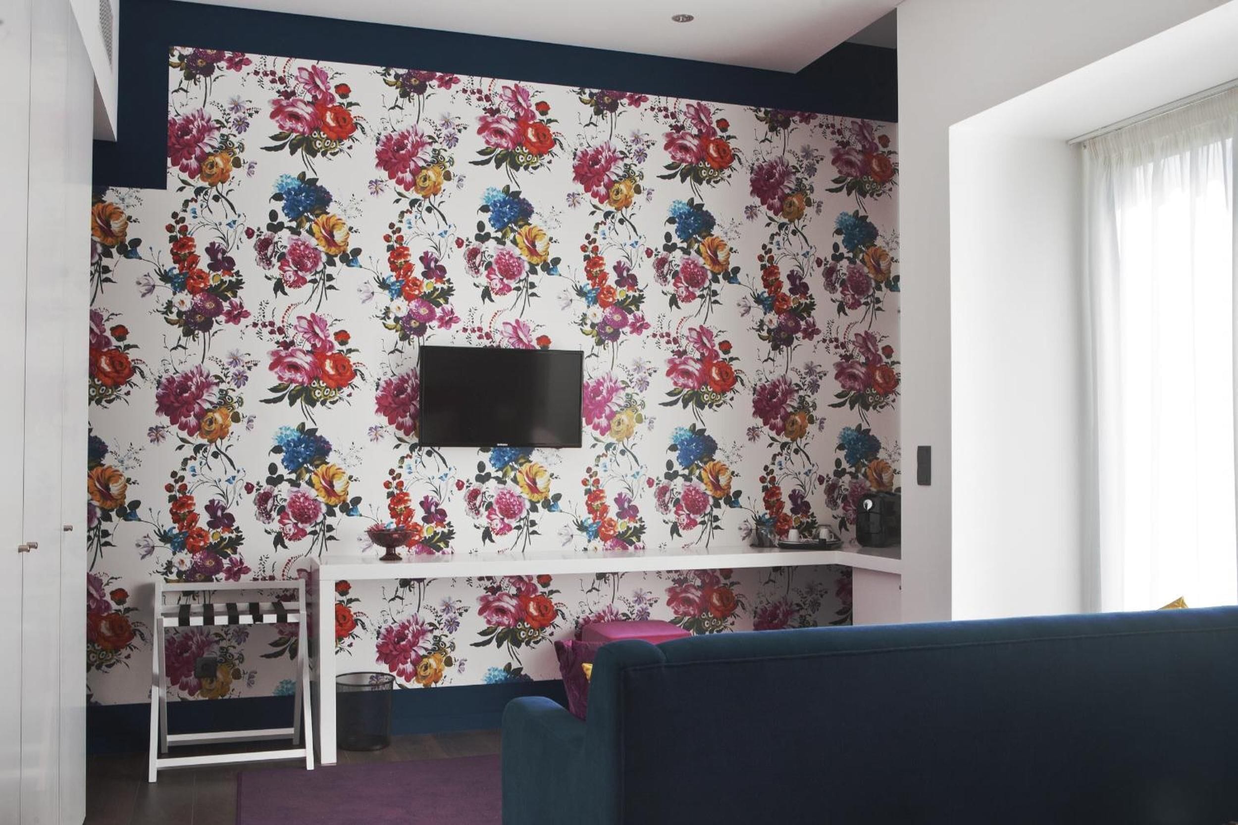 Un salon avec un papier peint à motifs floraux, un téléviseur à écran plat fixé au mur, un canapé bleu et une petite table blanche sur laquelle se trouve un panier.