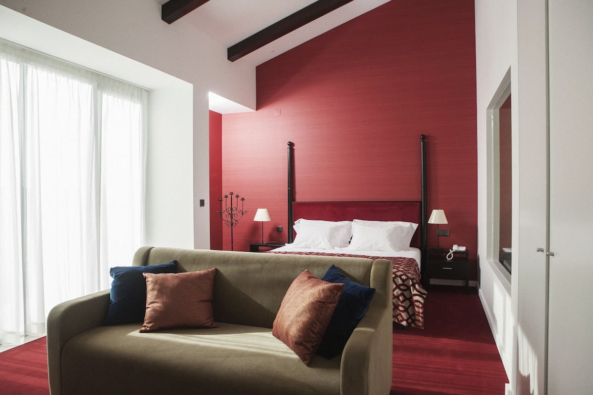 Habitación de hotel con pared de acento roja, cama con ropa blanca, sofá con varios cojines y mesita de noche con lámpara.