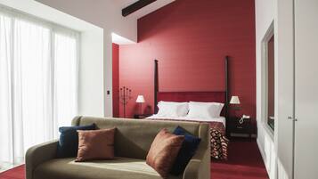 Habitación de hotel con pared de acento roja, cama con ropa blanca, sofá con varios cojines y mesita de noche con lámpara.