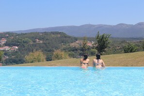 A swimming pool with two people in the water, surrounded by a hilly landscape with houses in the background. - Real Abadia Congress & SPA Hotel (Alcobaça)