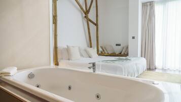 A modern bathroom with a large bathtub, a wooden structure on the wall, and a bed with white bedding in the background.