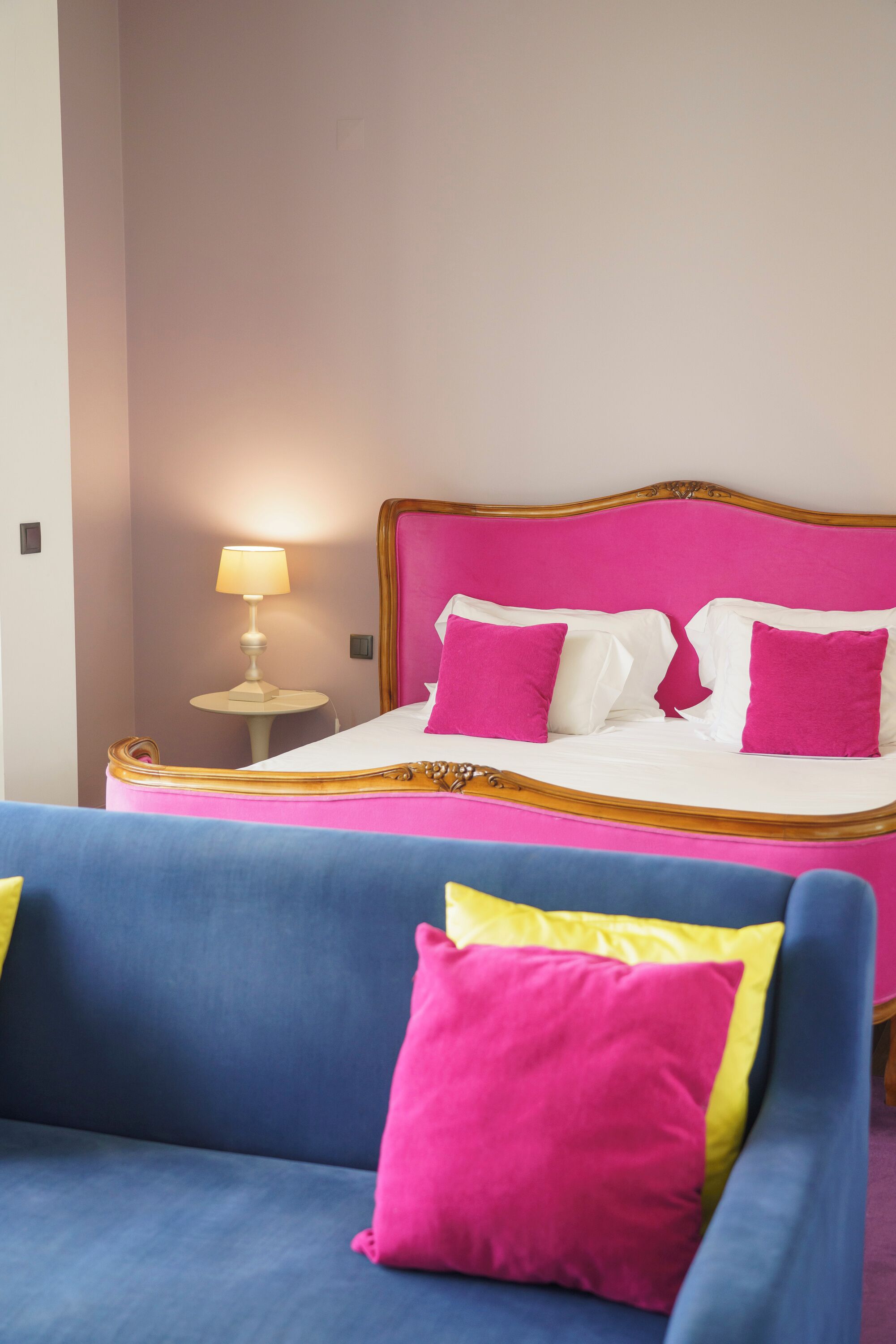A bed with a pink headboard and white bedding, a small bedside table with a lamp, and a blue sofa with a pink pillow.