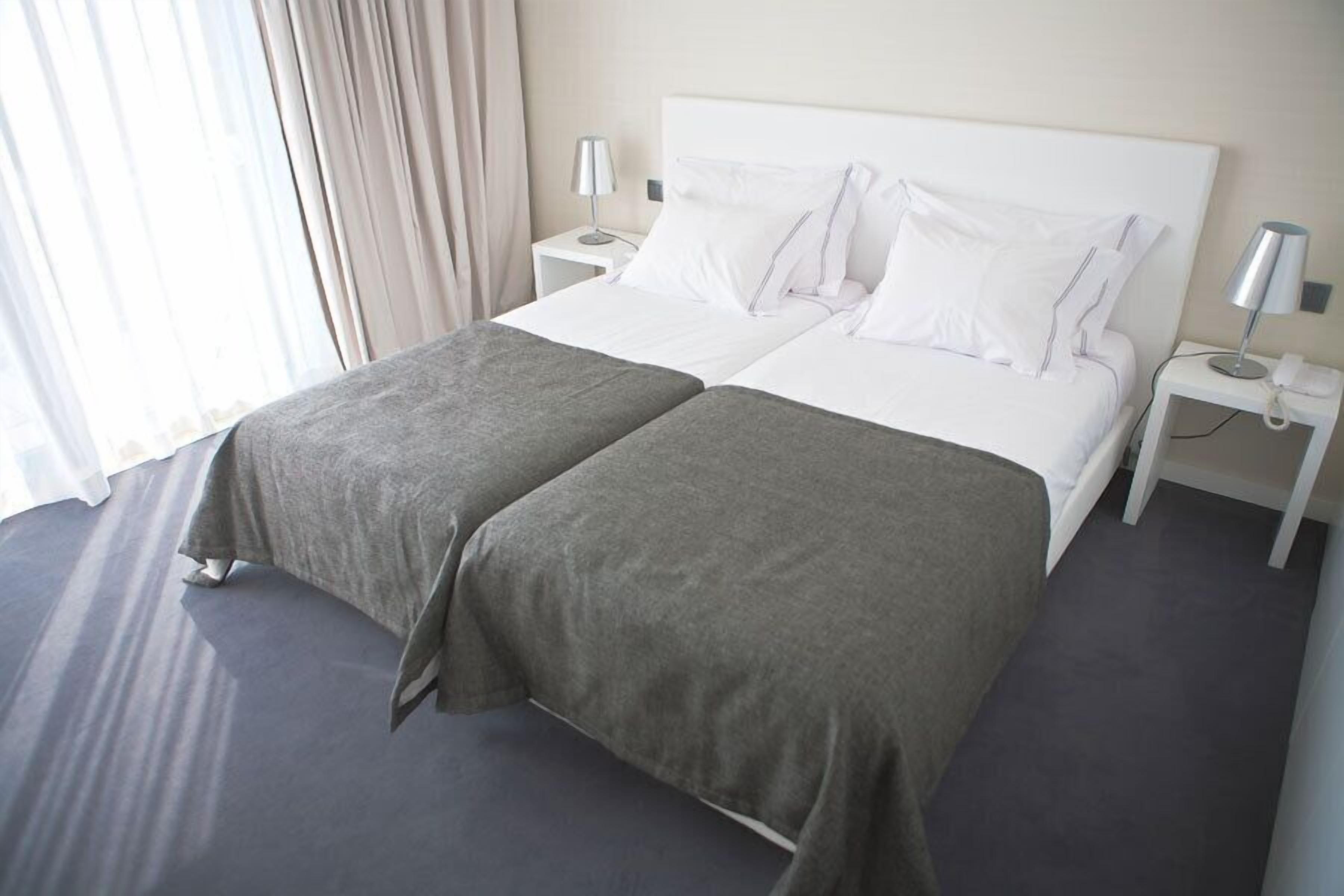 A double bed with white linens and a grey throw blanket, two bedside tables with lamps, and a window with sheer curtains.