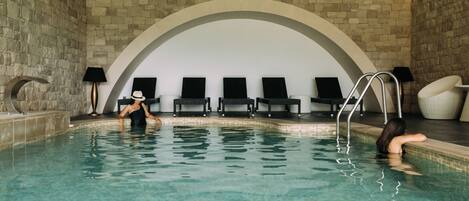 Una piscina interior con paredes de piedra, sillones reclinables y agua cristalina.