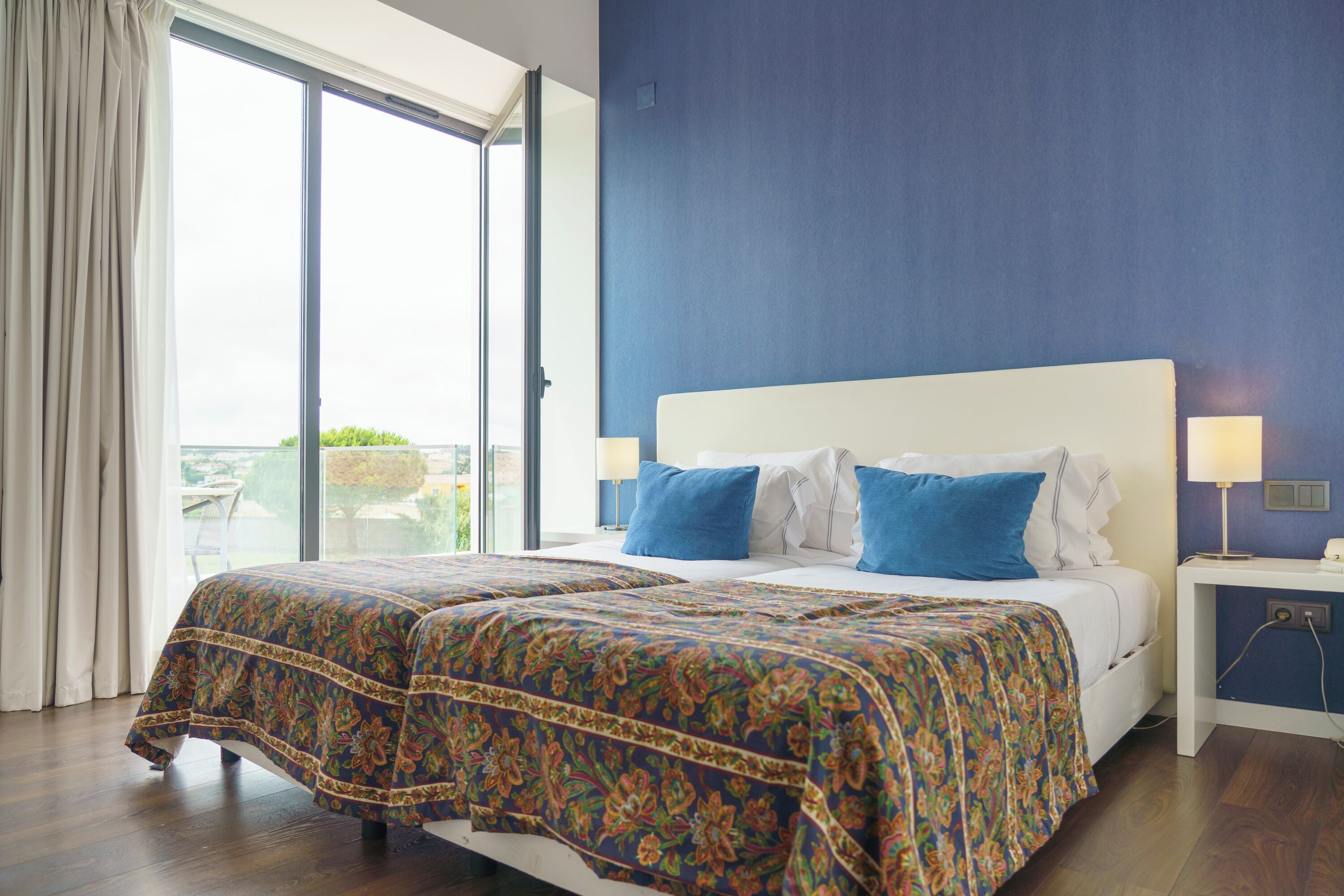 A double bed with floral bedding, a nightstand with a lamp, and a view through a sliding glass door.