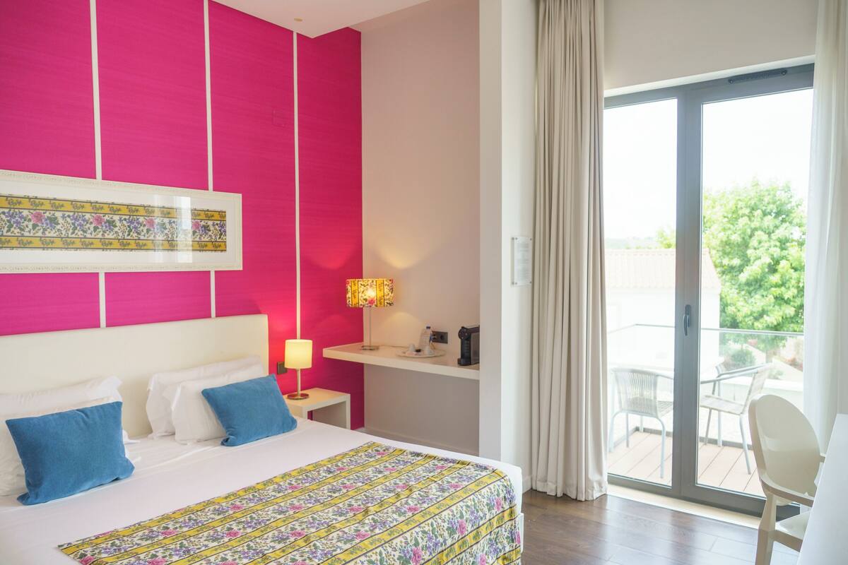 Habitación de hotel con cama, mesita de noche, una pequeña mesa, una silla, ventana con cortinas y balcón con mobiliario exterior.