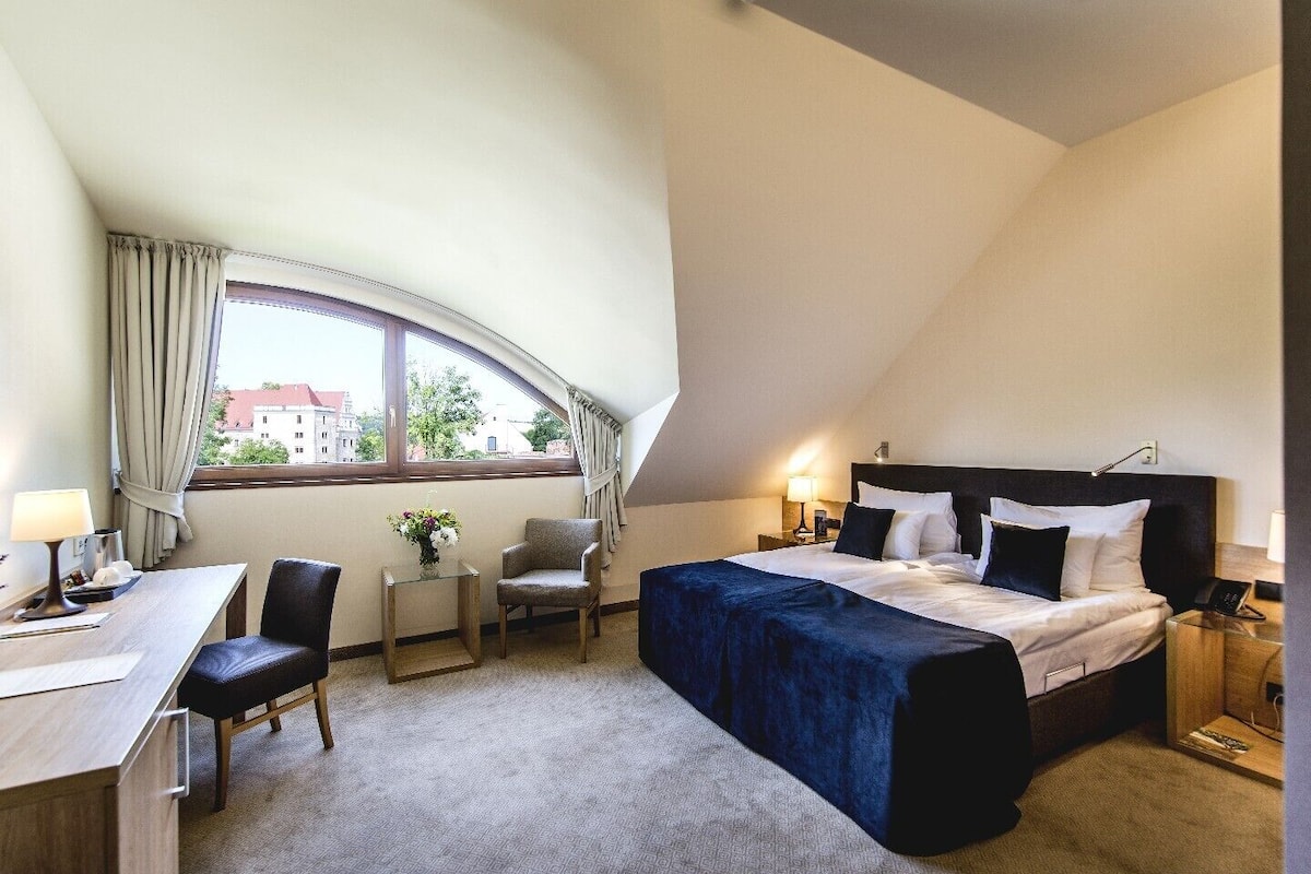 A hotel room with a curved ceiling, a bed with a dark headboard, a desk with a lamp, a chair, and a small table with a vase of flowers.