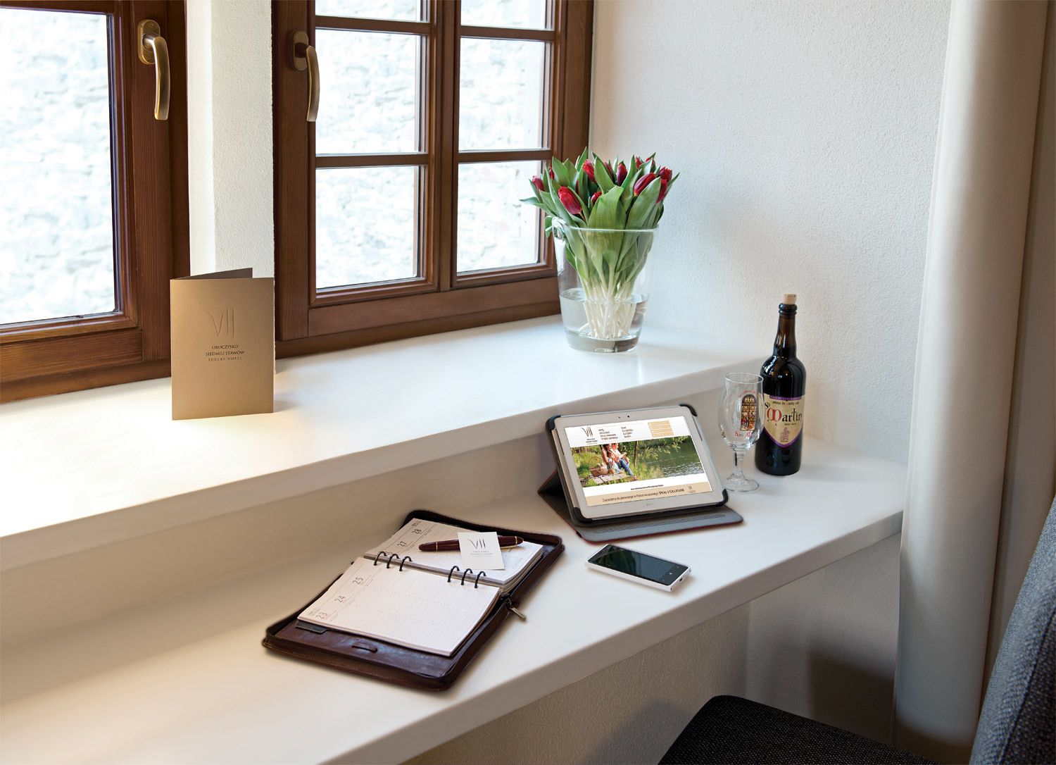 Ein Hotelzimmer mit Fenster, Schreibtisch mit Laptop, Tablet, Notizbuch, Stift, Smartphone, Weinflasche und Blumen in einer Vase.