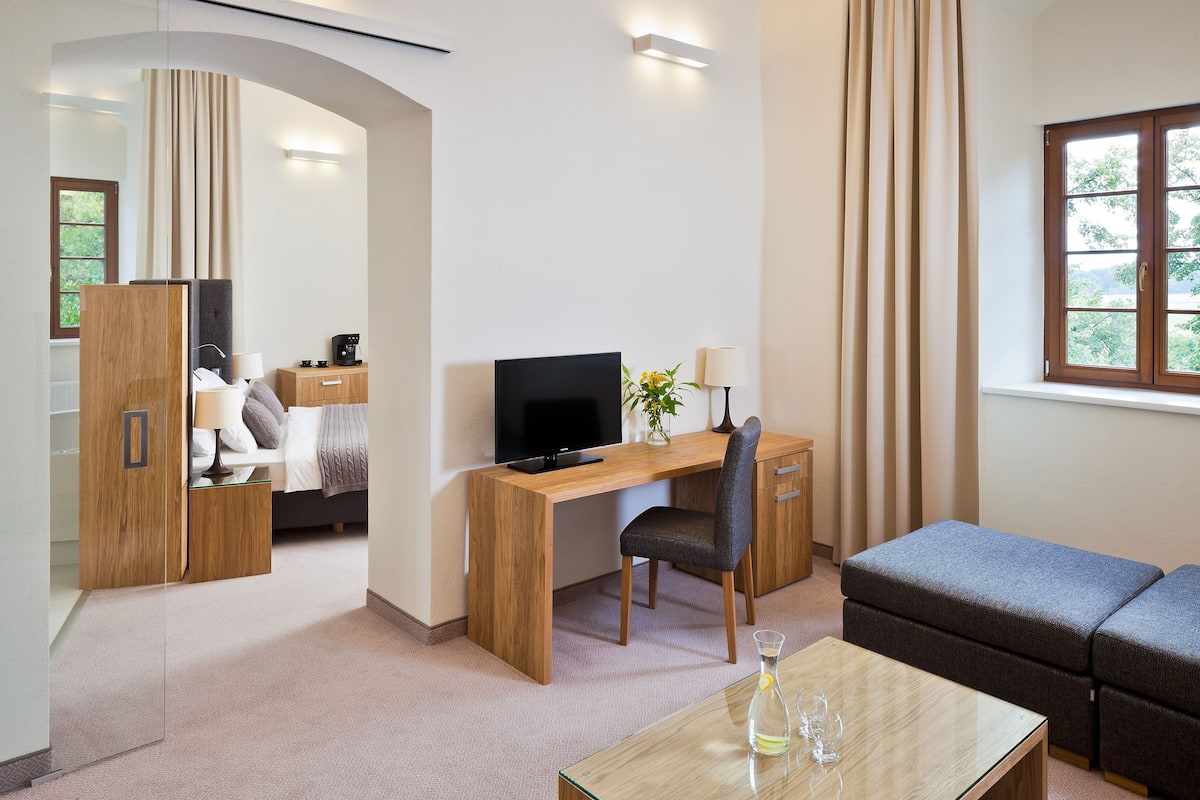 A modern hotel room with a wooden desk, a comfortable sofa, a glass coffee table, and a large window with curtains.