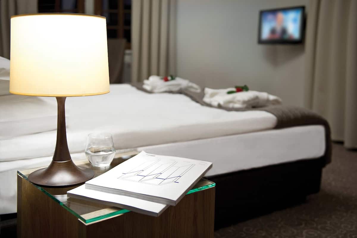 A hotel room with a bed, a bedside table with a lamp, a glass of water, and a document on the table.