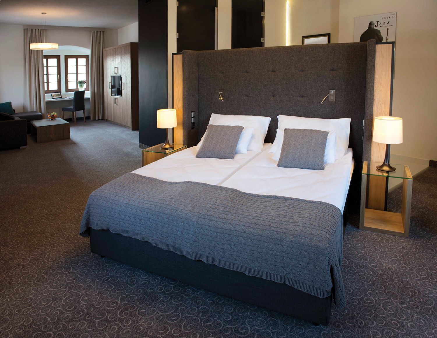 A hotel room with a large bed, two bedside lamps, a desk with a chair, and a view of the city through large windows.