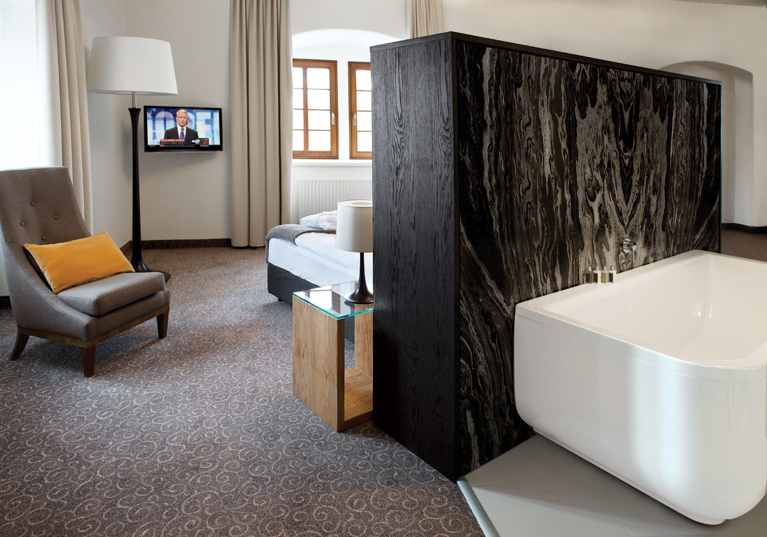 A modern hotel room with a large marble bathtub, a comfortable armchair with a yellow pillow, a television on the wall, and a bed with a white headboard.
