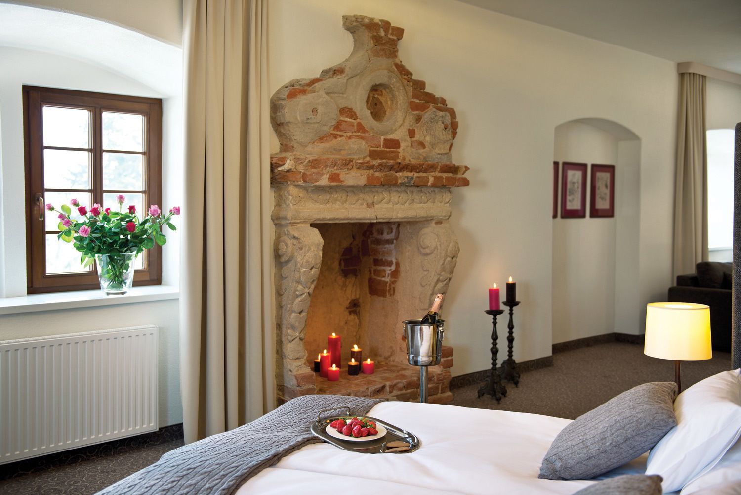Une chambre d’hôtel avec une cheminée en pierre, un lit avec des oreillers, une table de chevet sur laquelle se trouve une assiette de fraises, un vase de fleurs et une fenêtre avec des rideaux.