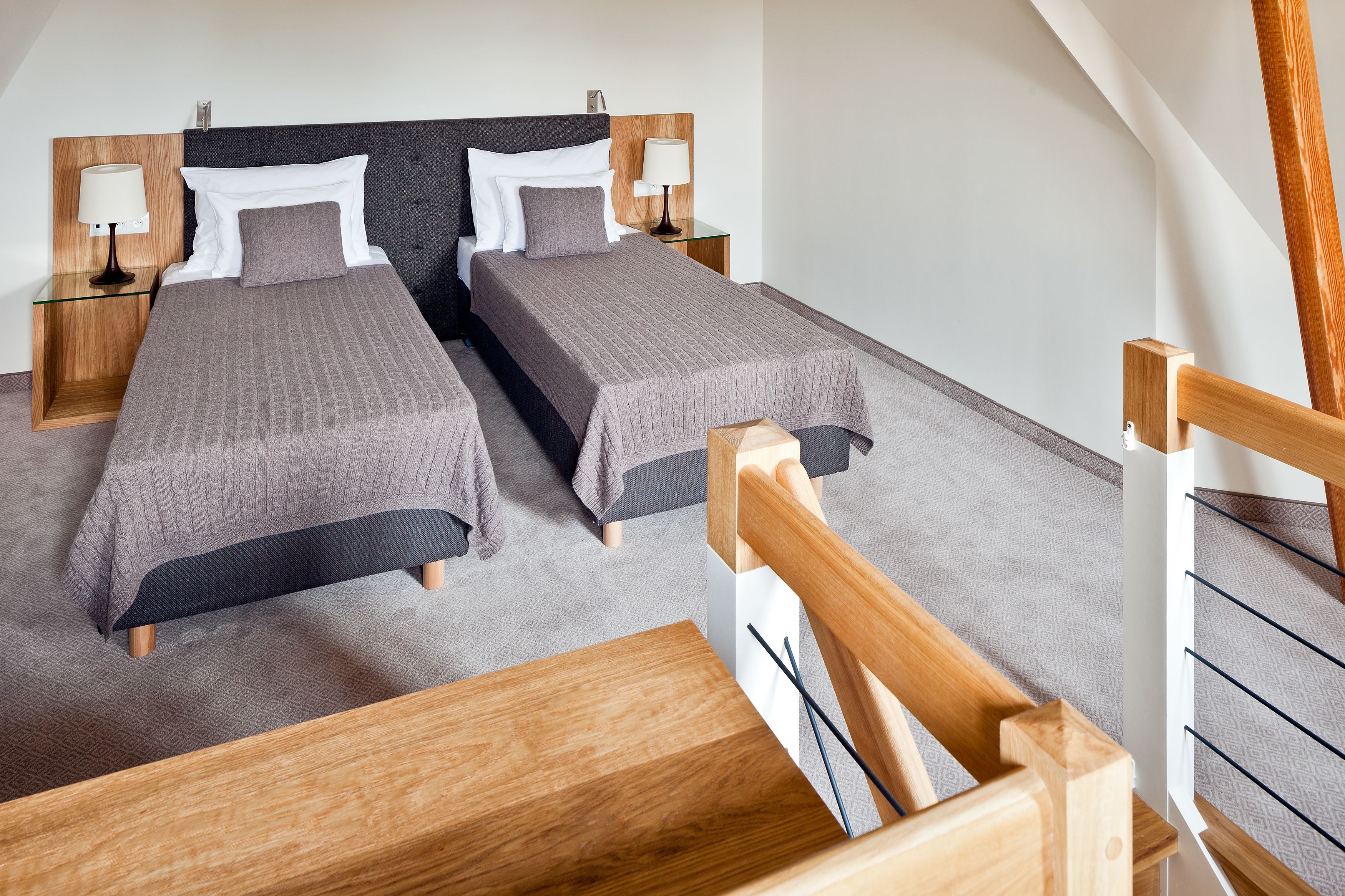 Ein modernes Hotelzimmer mit zwei Einzelbetten, einem Holzkopfstück, Nachttischen mit Lampen und einer Holztreppe.