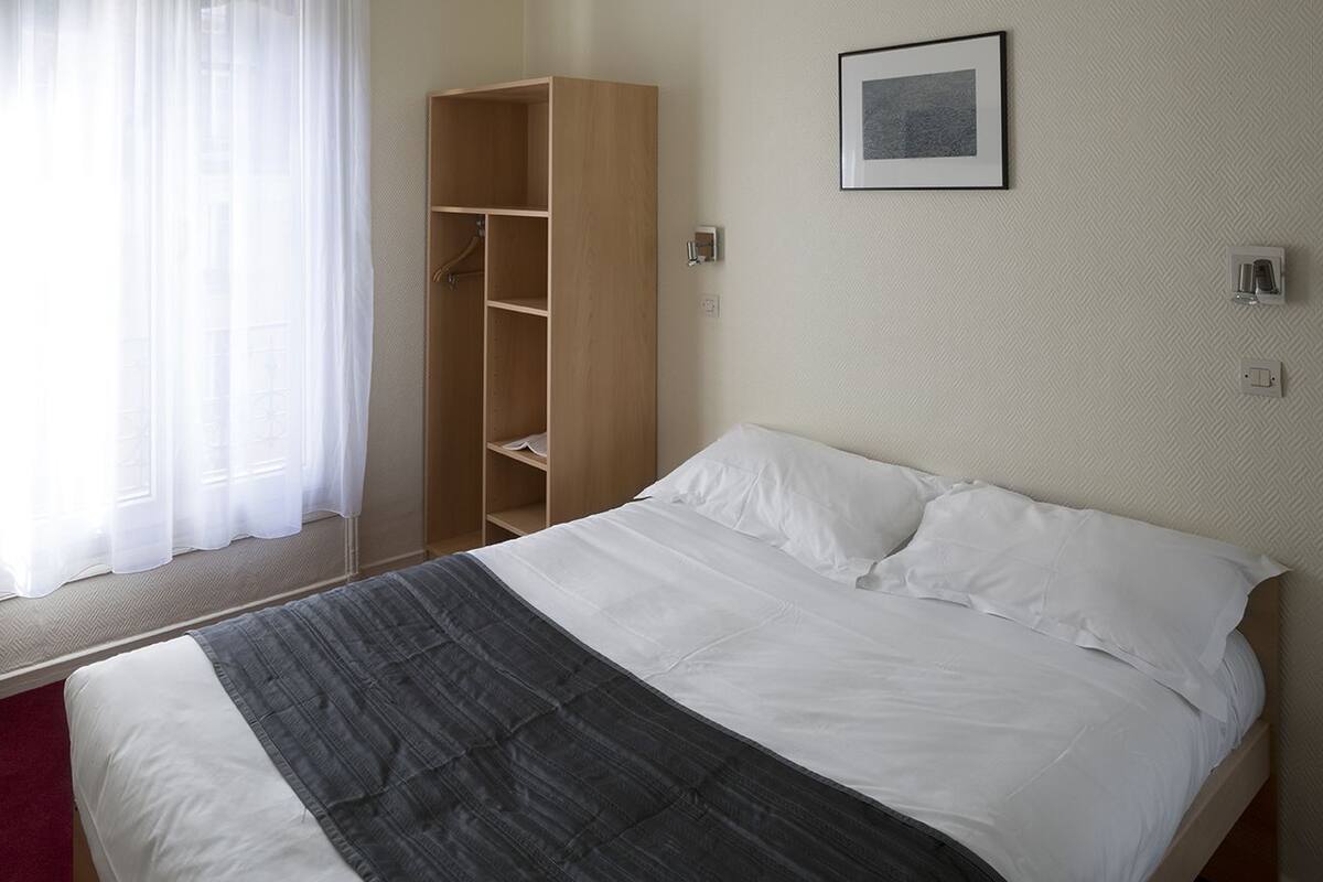 A bedroom with a bed, a wooden wardrobe, a window with sheer curtains, and a framed picture on the wall.