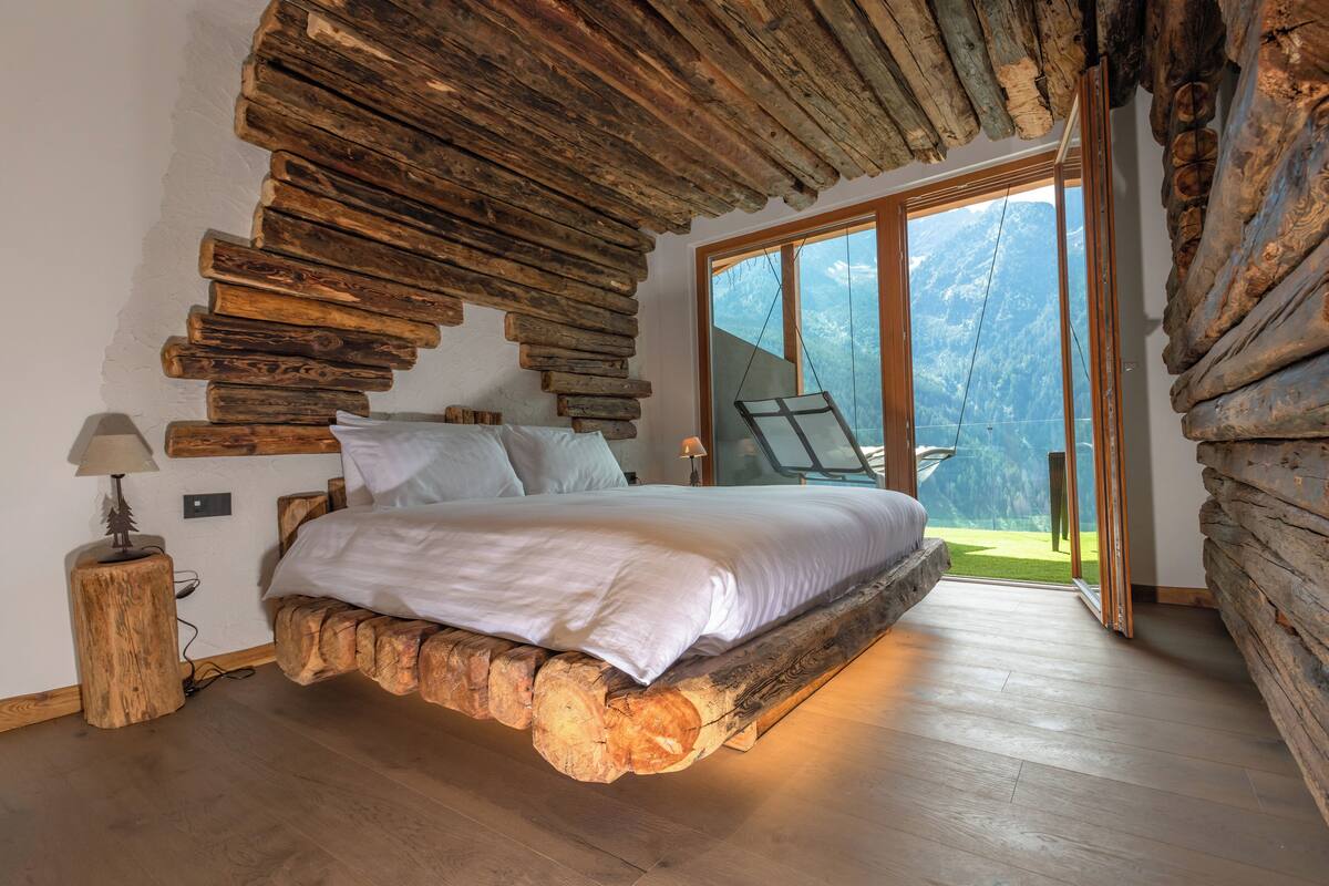 A bedroom with a bed on a log base, wooden walls, a nightstand, and a view of a mountain through a glass door.