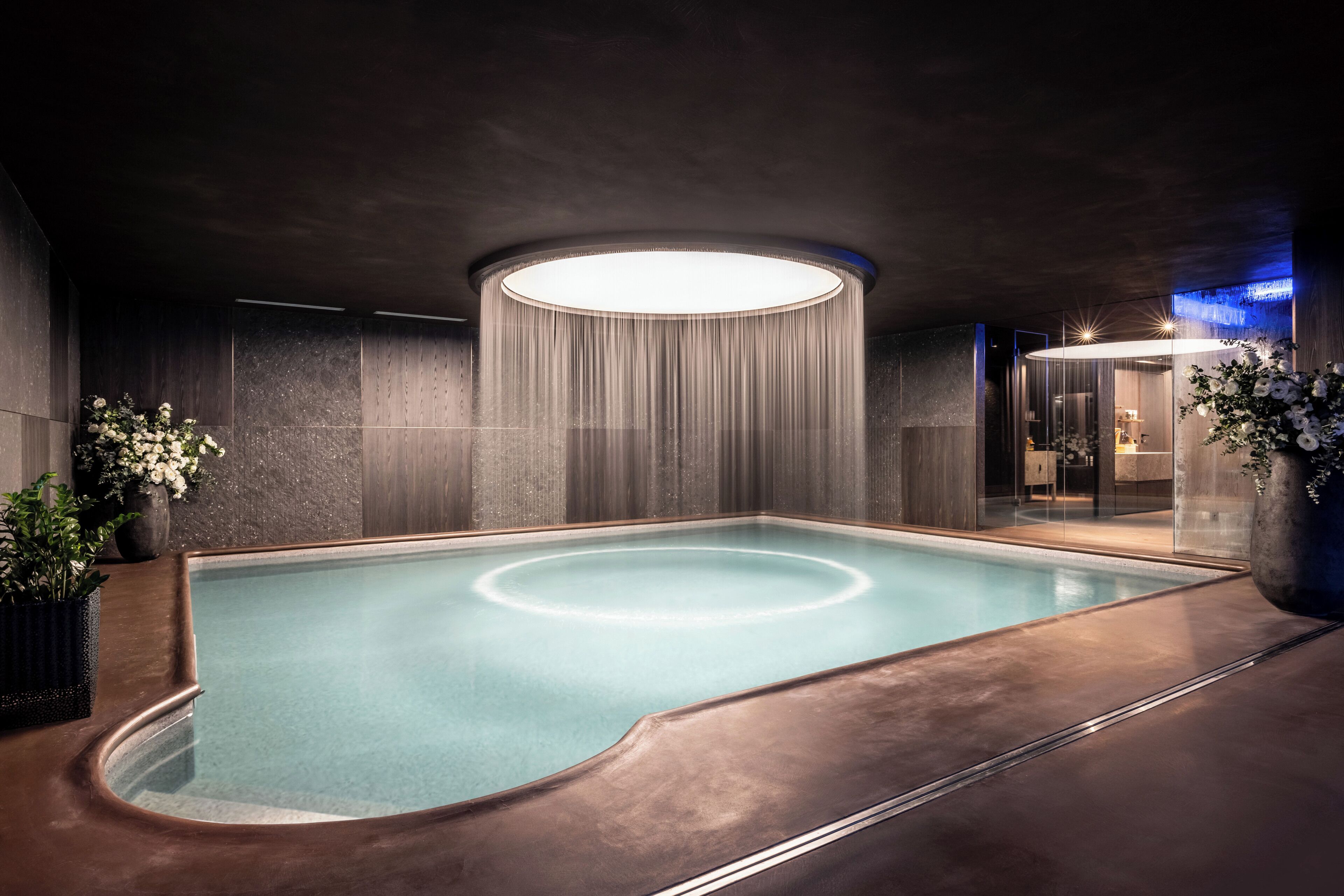 A modern indoor pool with a circular light fixture above, surrounded by dark walls and potted plants.
