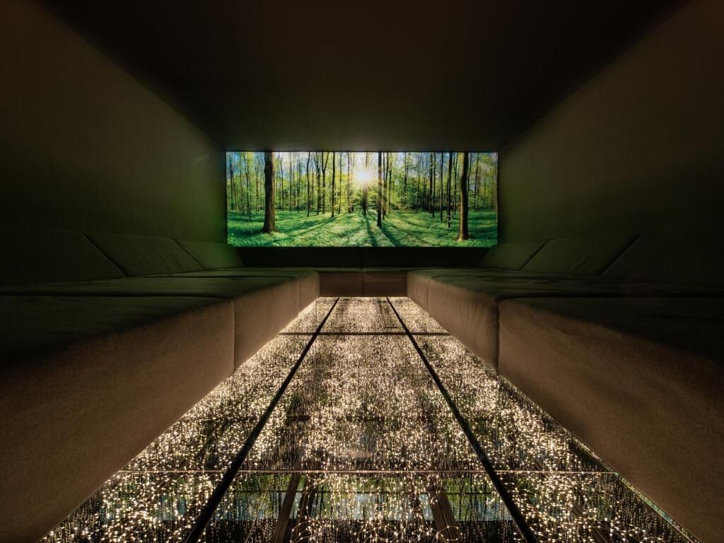 A room with a large flat-screen TV on the wall displaying a forest scene. The room has a tiled floor and a low ceiling.