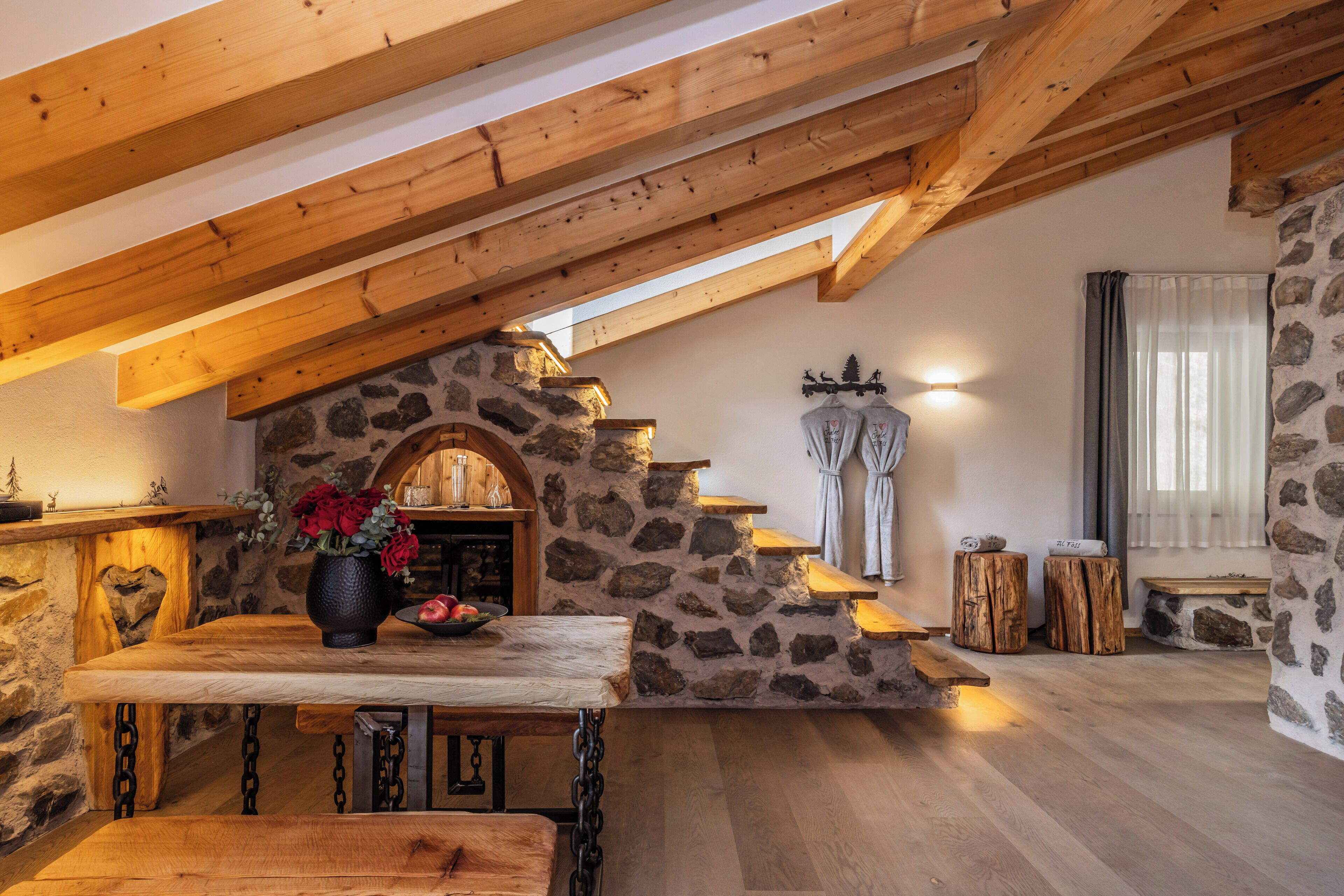 Un interior rústico con chimenea de piedra, vigas de madera y una mesa de madera con un jarrón de flores.