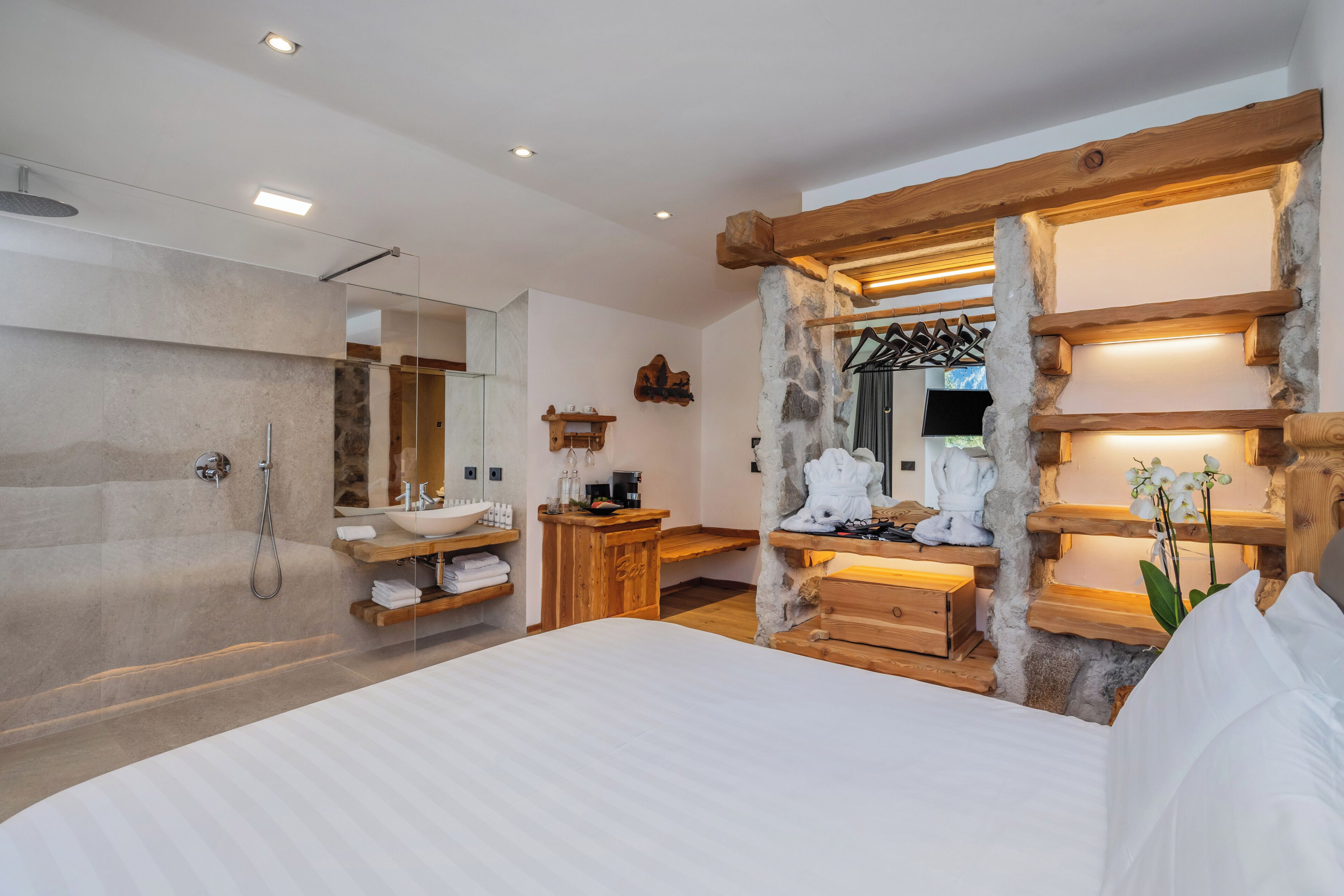 Un dormitorio moderno con una cama grande, pared de piedra, vigas de madera, un escritorio de madera, tocador con lavabo y ducha.