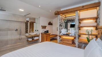Un dormitorio moderno con una cama grande, pared de piedra, vigas de madera, un escritorio de madera, tocador con lavabo y ducha.