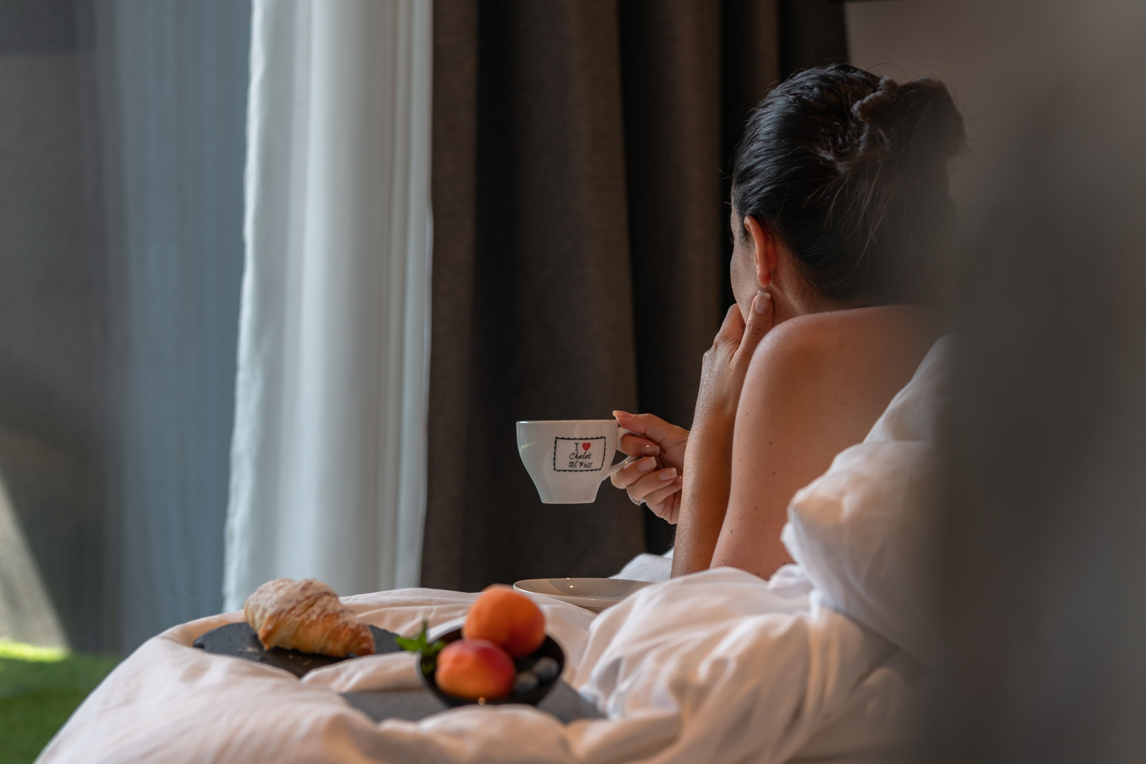Una persona seduta su un letto con una tazza e un piatto di cibo, con un cornetto e della frutta.