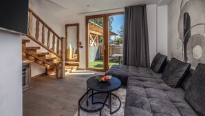 A modern living room with a wooden staircase, a grey sofa, and a dining area visible through a glass door.