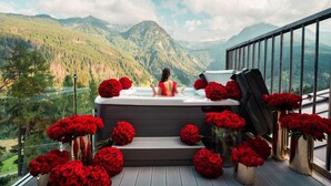 Tinaja de hidromasaje con una mujer adentro, rodeada de rosas rojas, con vista a un paisaje montañoso.