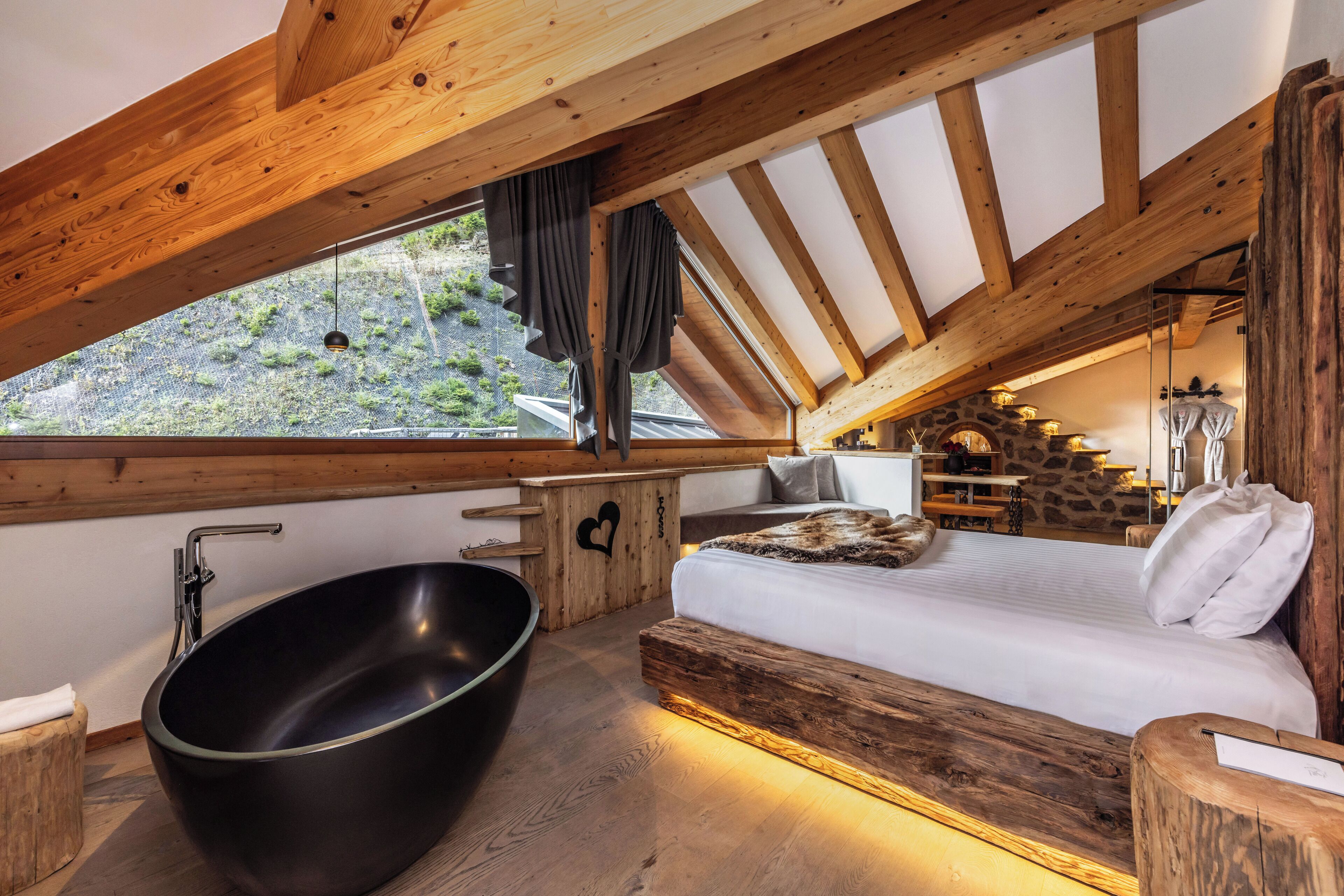 A bedroom with a large bathtub, wooden beams, a bed with white bedding, and a view of a mountainous landscape.
