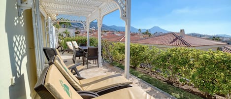 A covered outdoor seating area with lounge chairs and a table, overlooking a landscaped garden and a view of buildings and mountains.