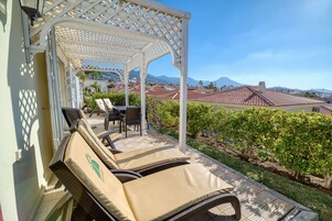 Un espacio al aire libre cubierto con sillones y una mesa, con vistas a un jardín ajardinado y edificios y montañas.