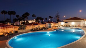 A swimming pool with lounge chairs and umbrellas, surrounded by palm trees and buildings at dusk. - Ona Sueño Azul (Adeje)