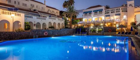A swimming pool with a clear blue water surface, surrounded by a stone wall, and adjacent to a building with multiple balconies and outdoor seating areas.